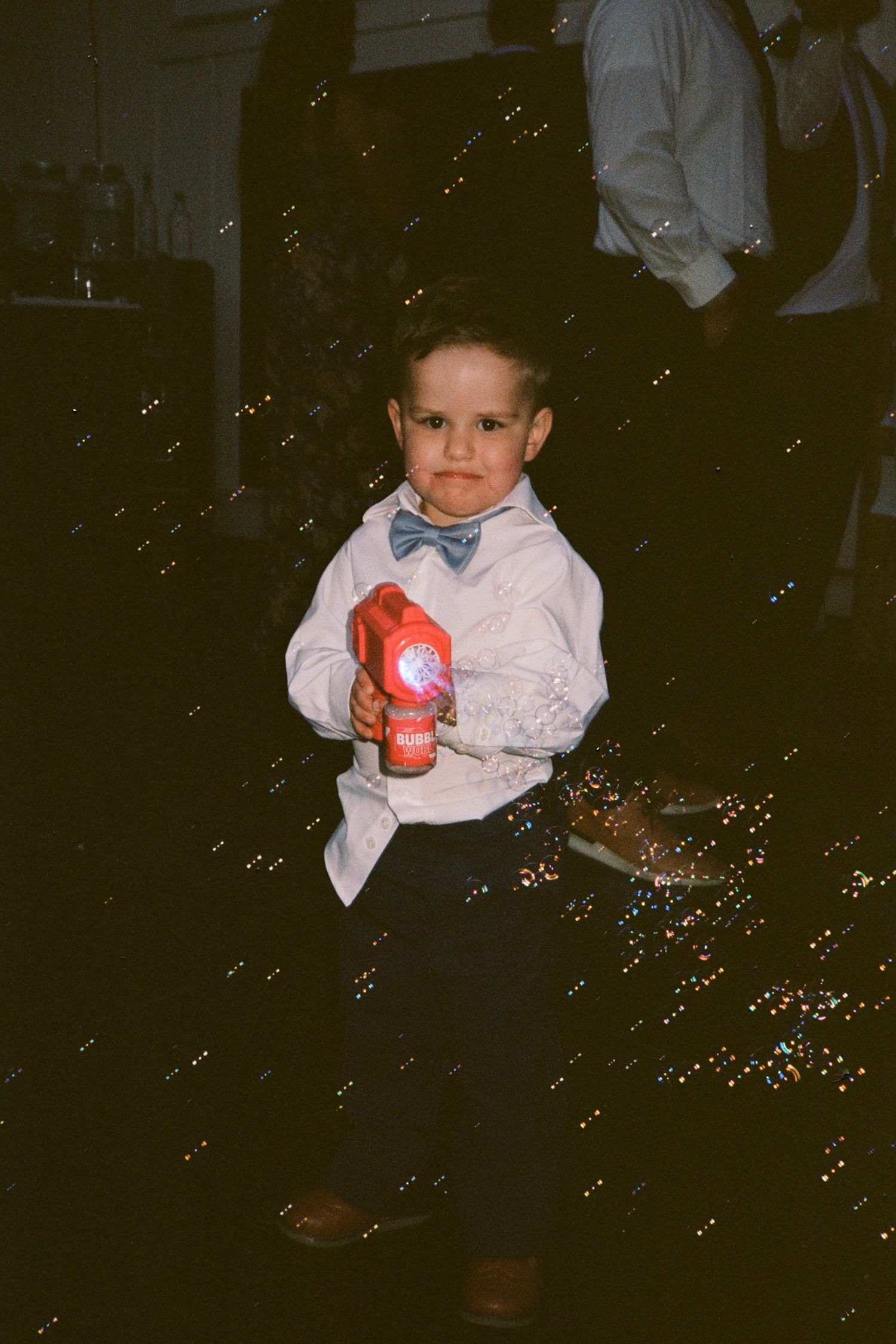 Adorable young boy in white shirt and bow tie holds toy at wedding reception with bubbles.