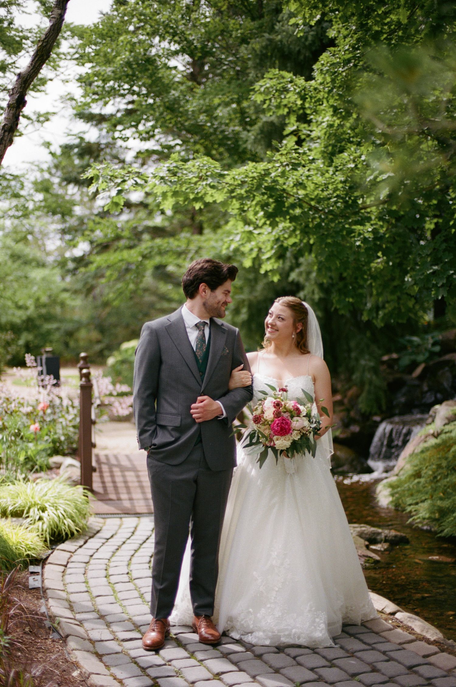 Bride and groom walk hand-in-hand on a cobblestone path in a lush garden, she holds a pink bouquet.