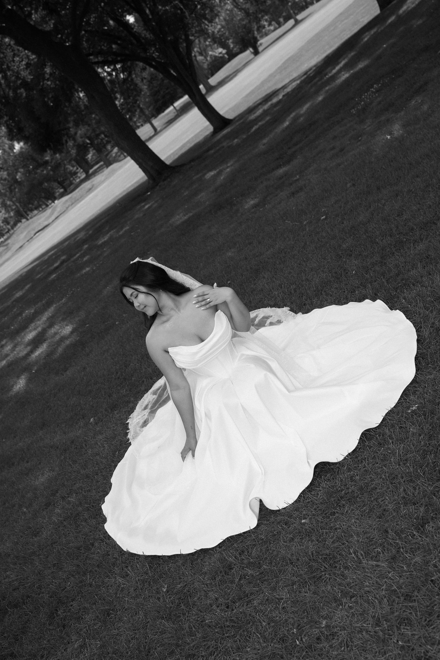 Bride in flowing white wedding gown sitting gracefully on grass in black and white outdoor photo.