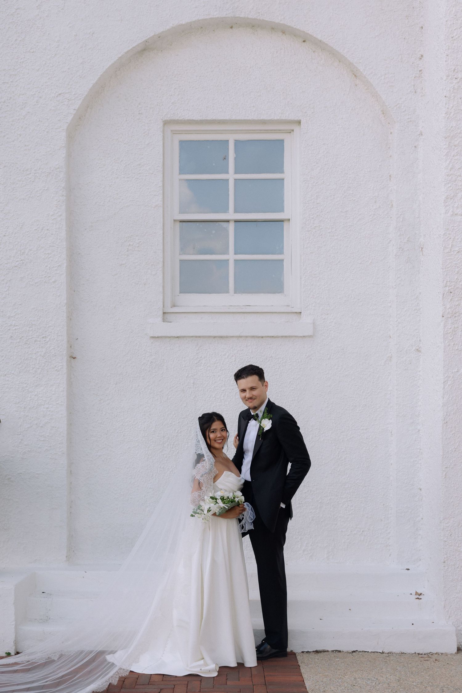 Smiling bride and groom posing together against a white arched wall with bouquet on wedding day.