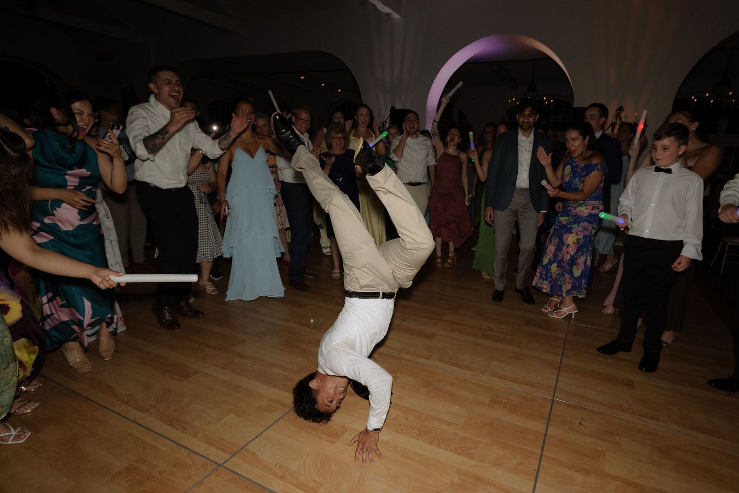 Wedding guest performing an impressive breakdancing headstand on the dance floor surrounded by cheering wedding guests.