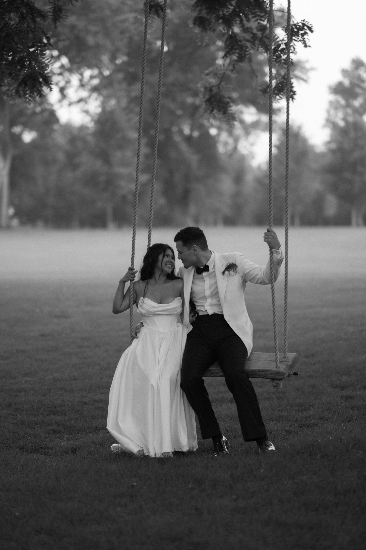 Black and white photo of bride and groom sitting together on a rope swing in a lush green park.