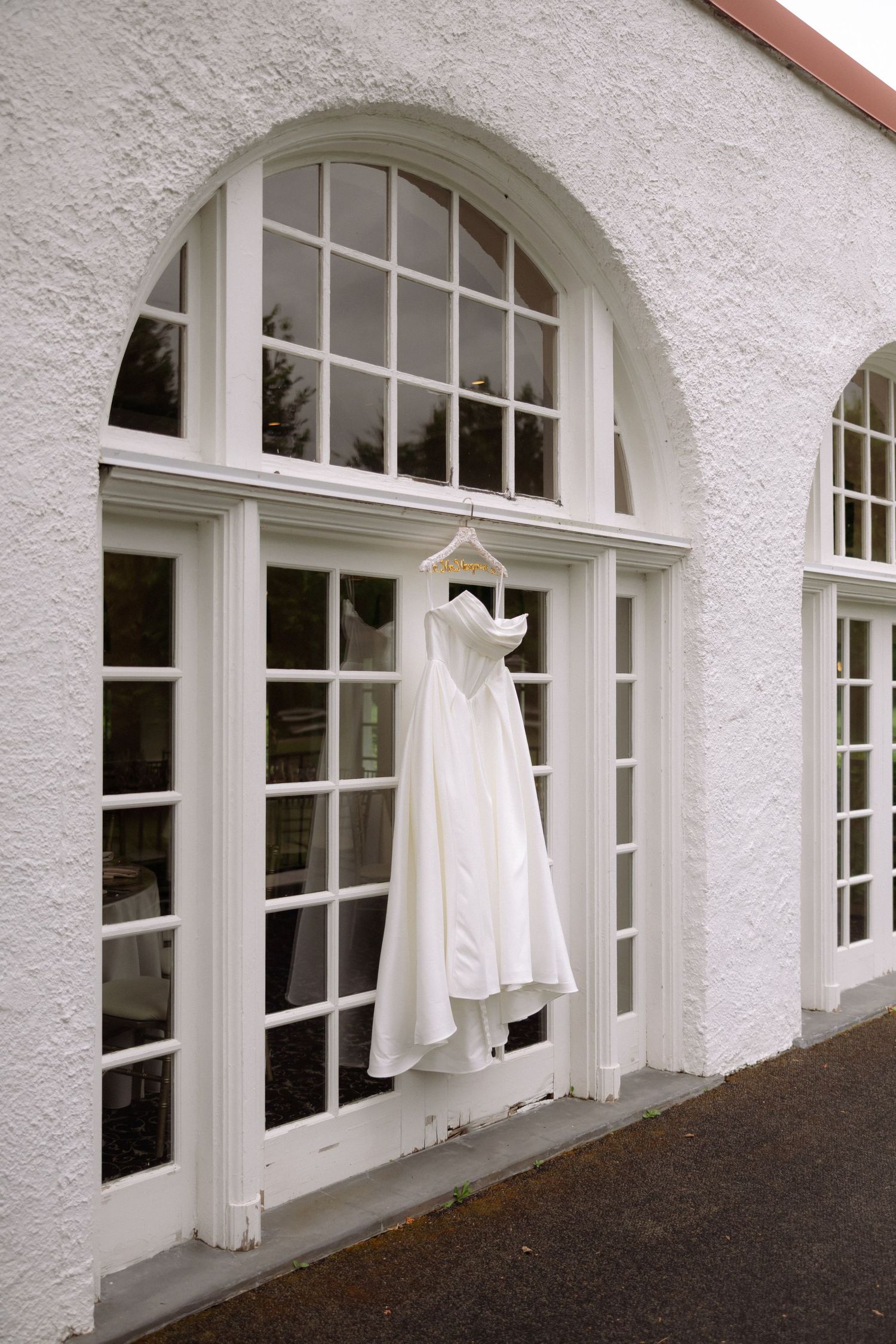 White wedding dress hanging on an arched French window of a white stucco building exterior.