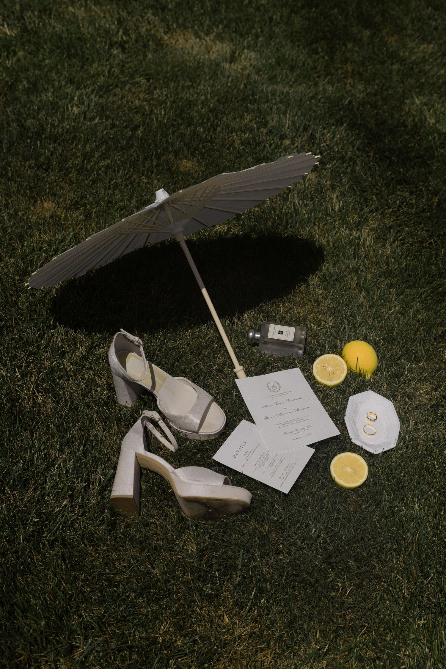 Wedding flat lay with heels, invitations, lemons, and perfume on grass under a black parasol umbrella.