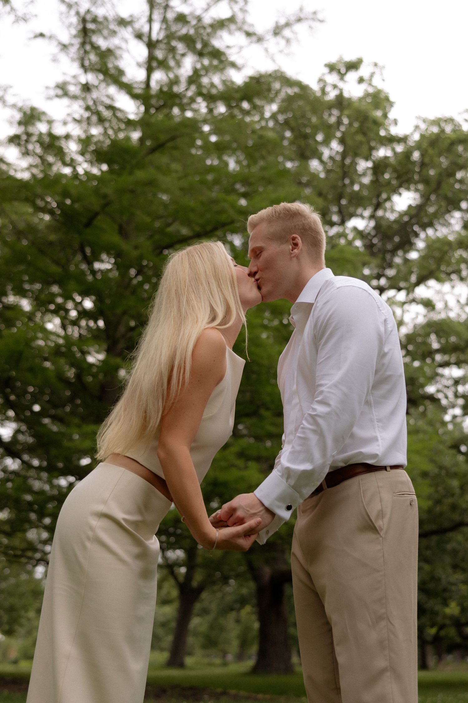 Romantic couple sharing a kiss outdoors in a green park, holding hands in coordinating neutral outfits.
