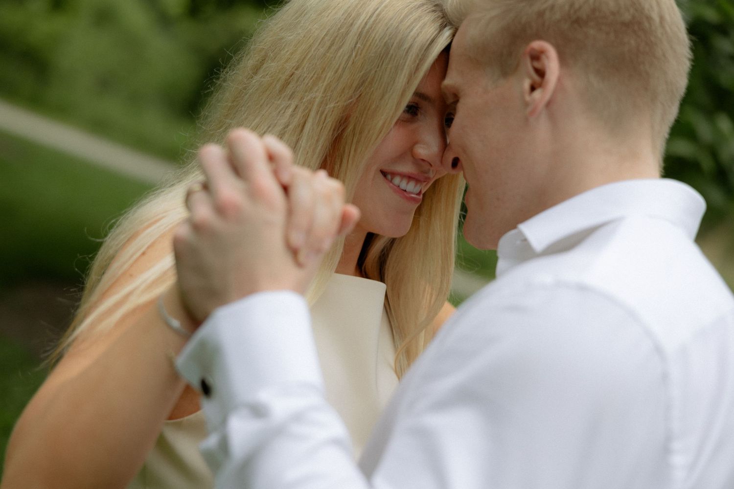 A smiling blonde woman and a man in a white shirt hold hands and dance closely together outdoors.