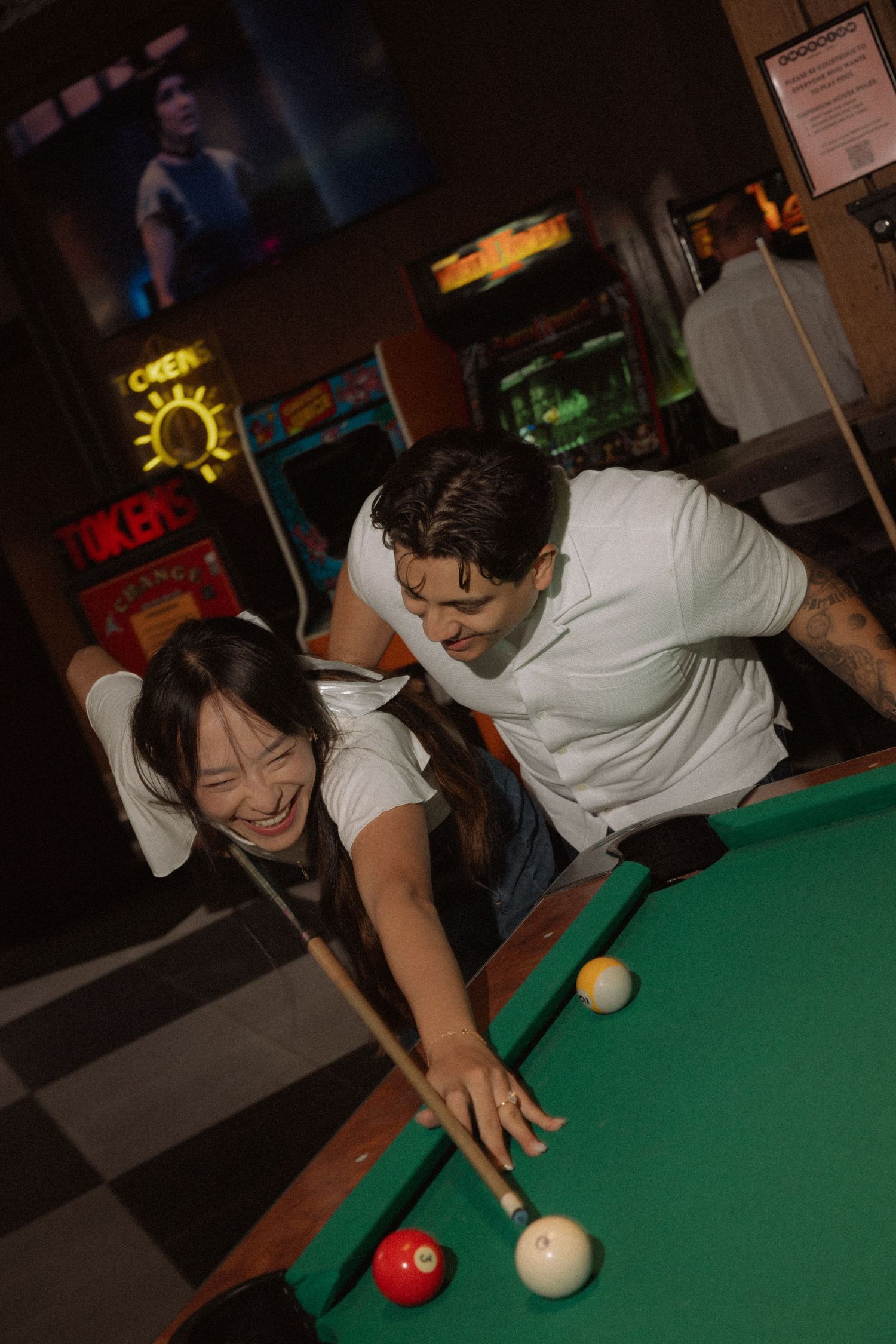 Couple laughing together while playing pool at a bar arcade.