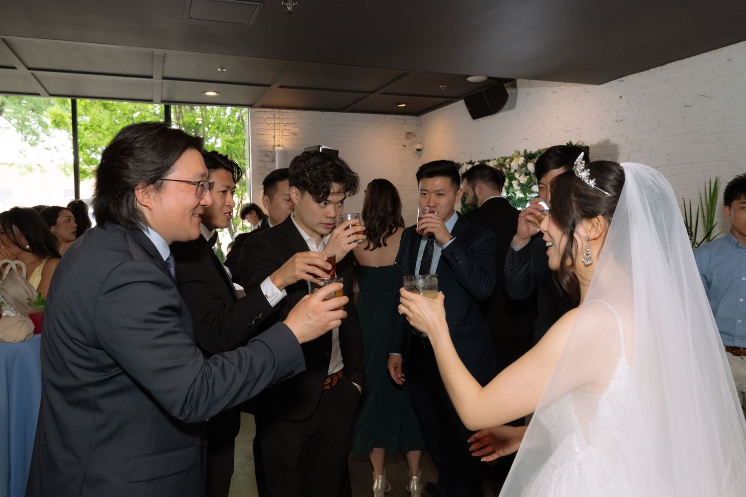 Wedding guests raising champagne glasses in a toast with a bride in a white veil at an elegant indoor reception.