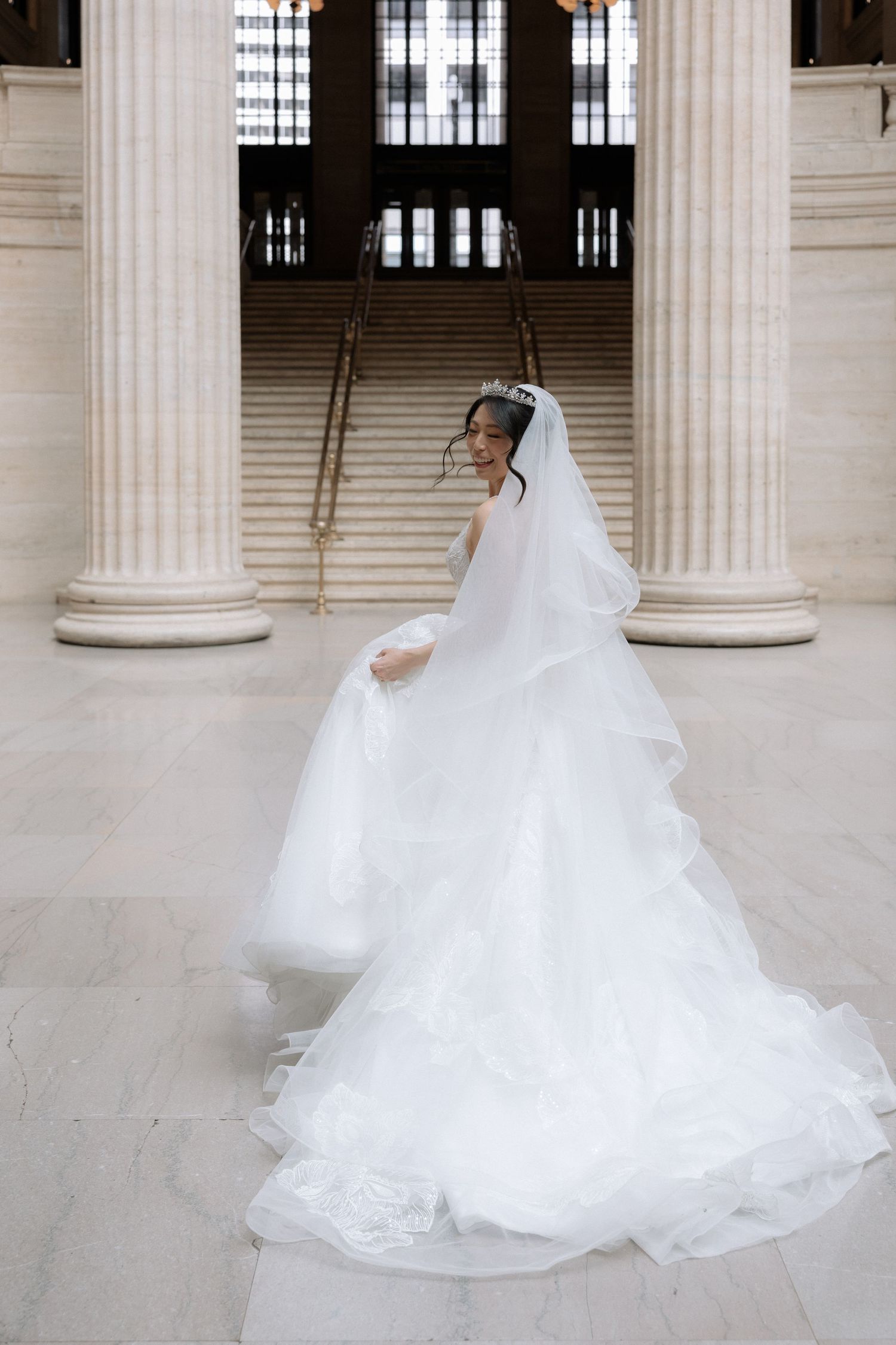 Bride in flowing white ball gown and veil twirls before grand columns at elegant wedding venue.