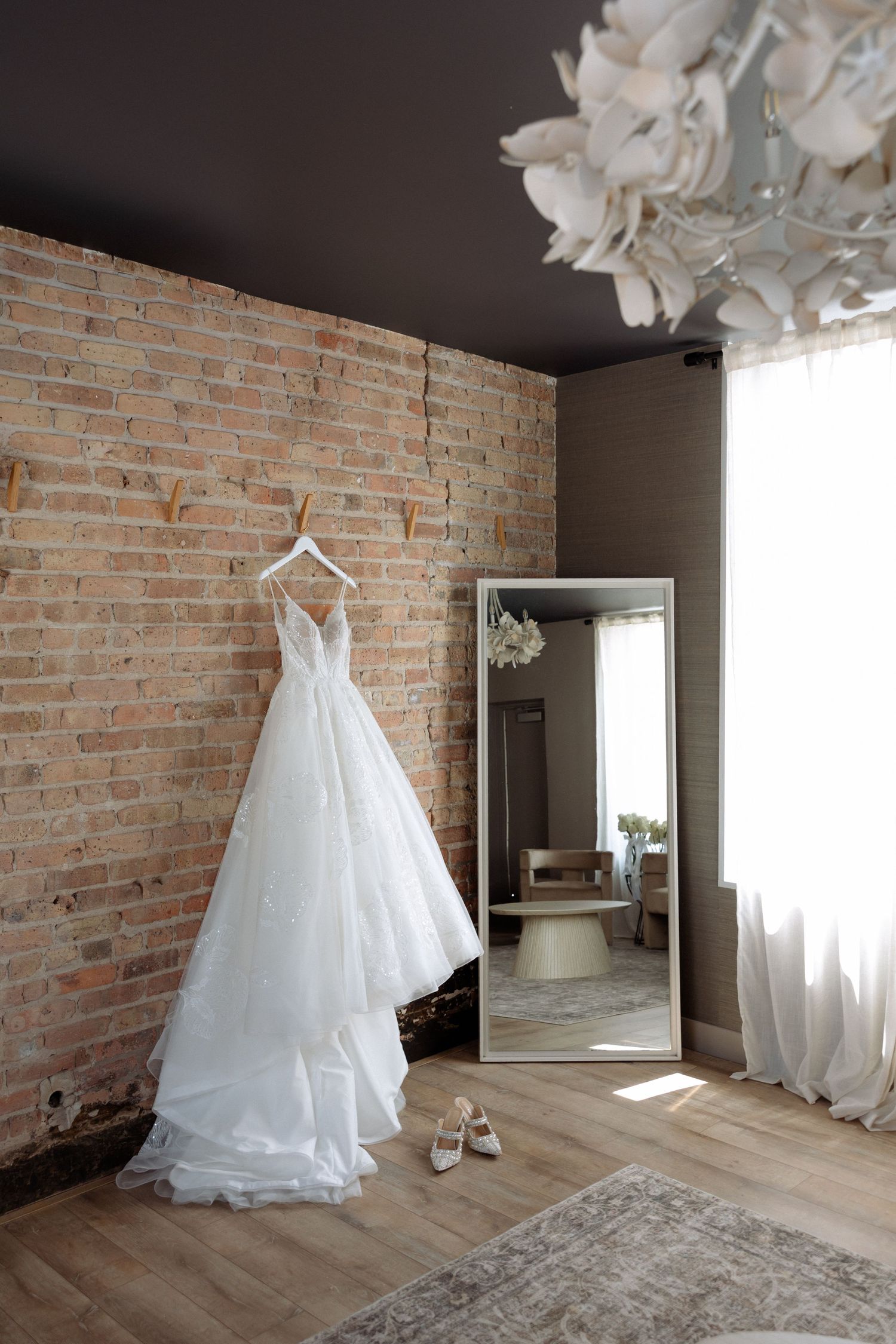 White wedding dress hanging on brick wall beside floor mirror with heels below in elegant bridal suite.