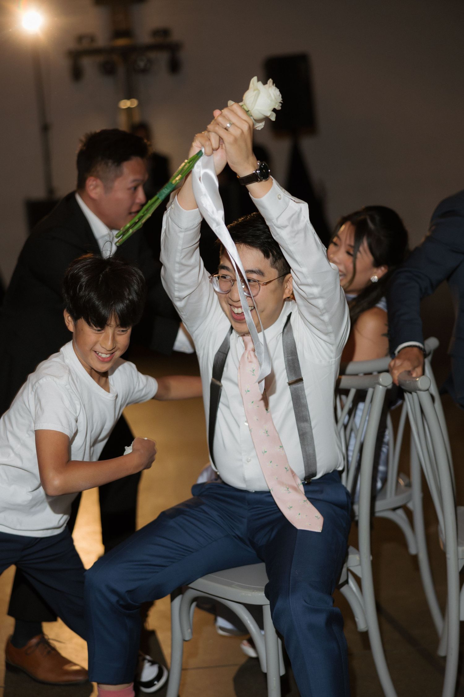 Man triumphantly raises white flower while seated at lively wedding reception game activity.