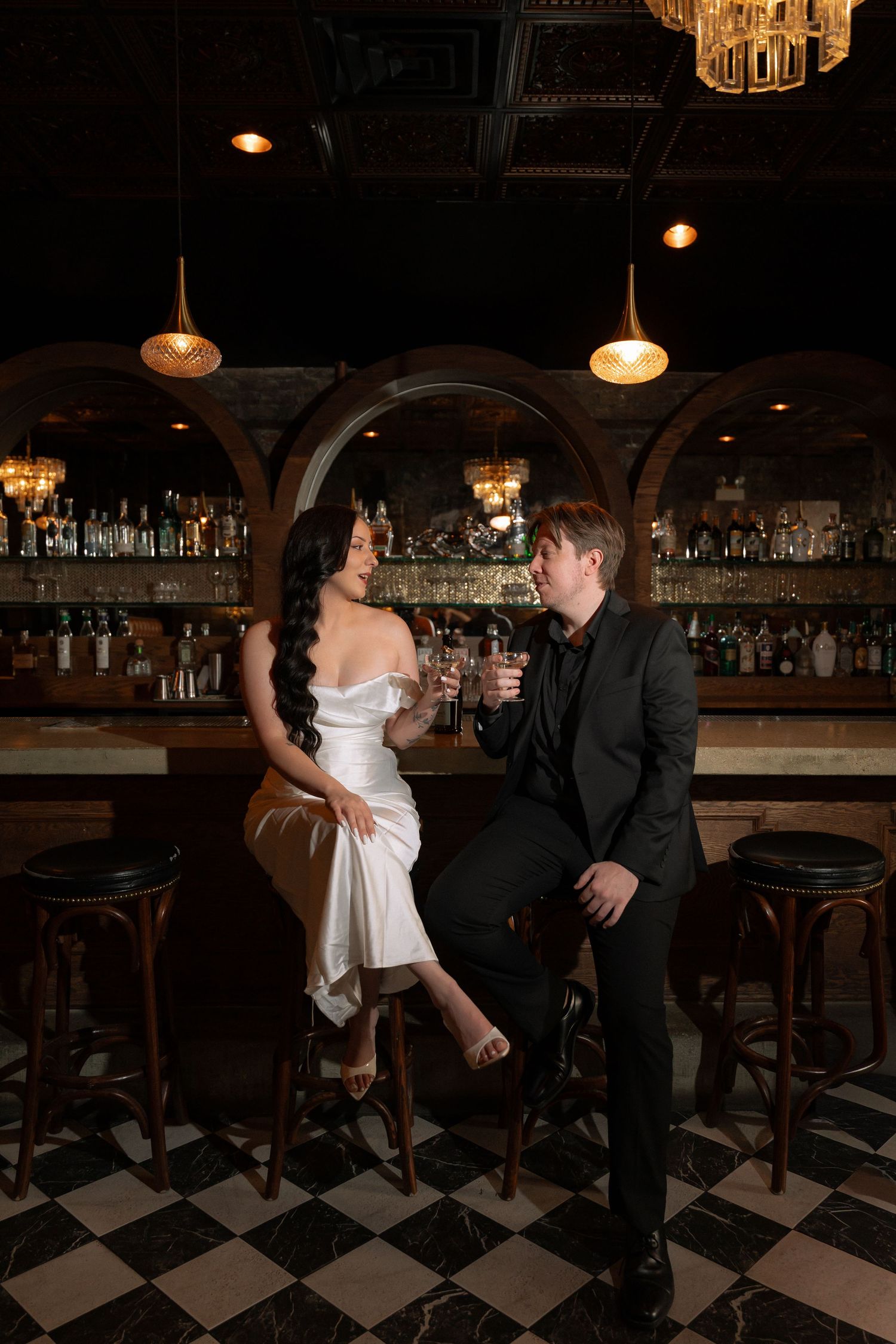 Bride in white gown and groom in black suit share drinks at an elegant dark bar with checkered floors.