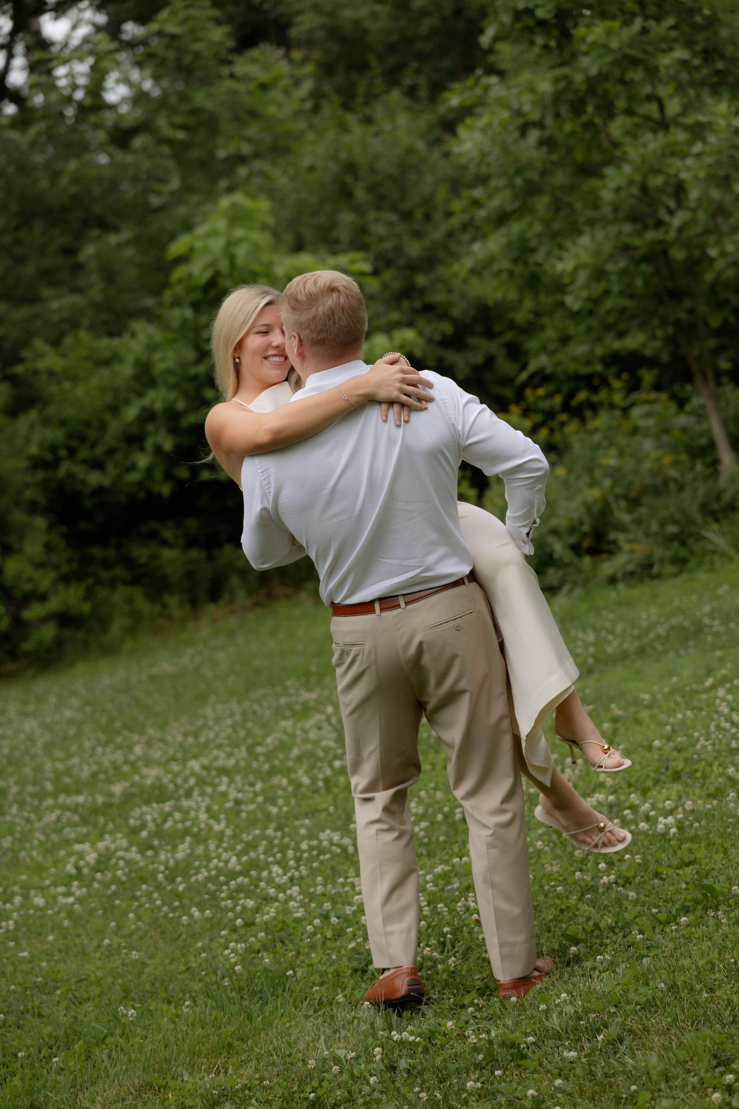 Man lifting and spinning smiling blonde woman in green park during romantic outdoor couples photo session.