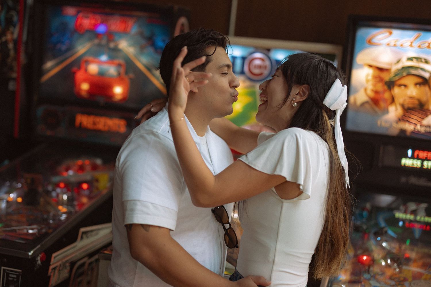Couple sharing a kiss in front of colorful pinball machines in an arcade, both wearing white outfits.