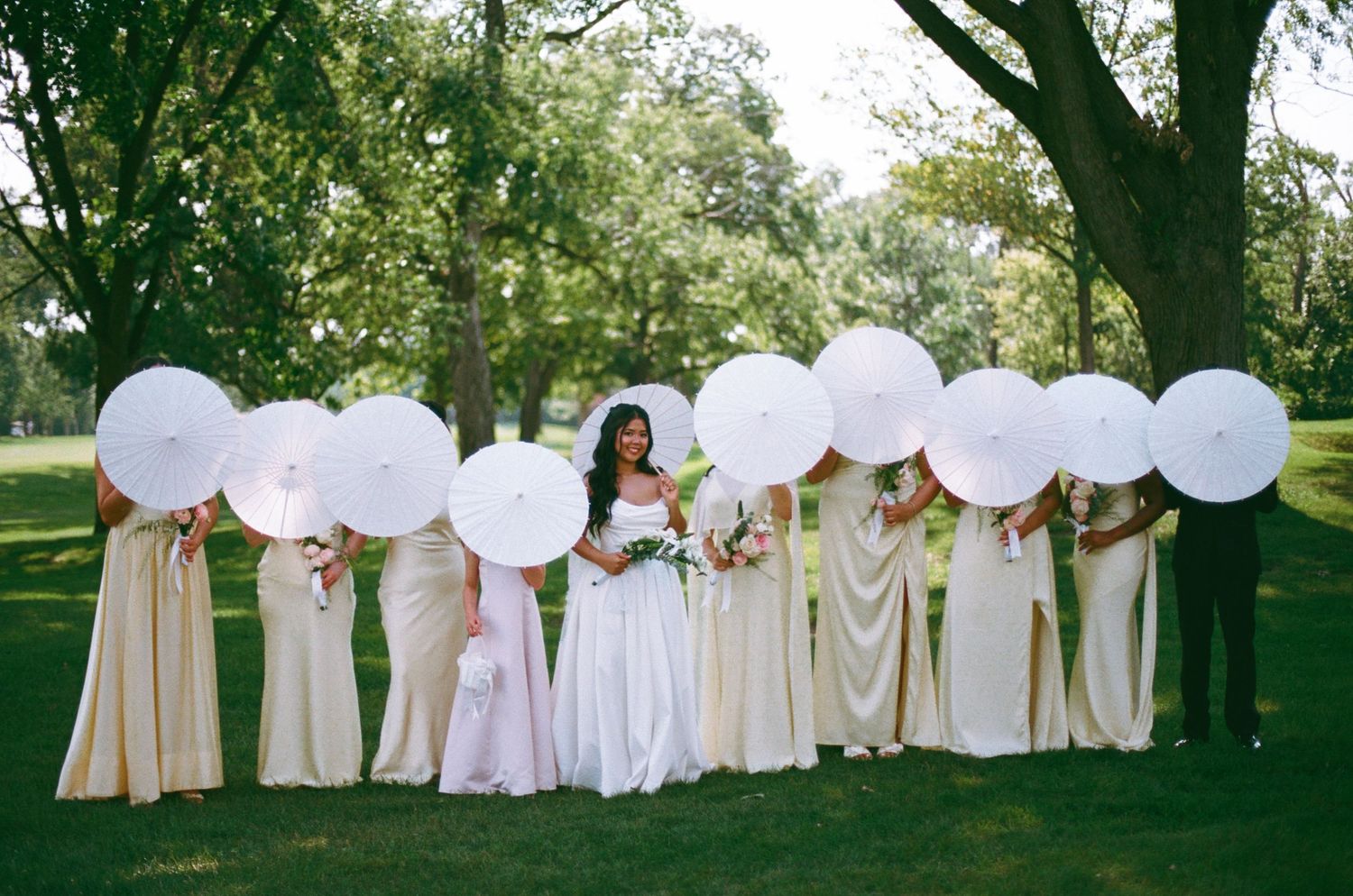 Bride and bridesmaids in ivory gowns holding large white parasols outdoors under green trees on a sunny wedding day.