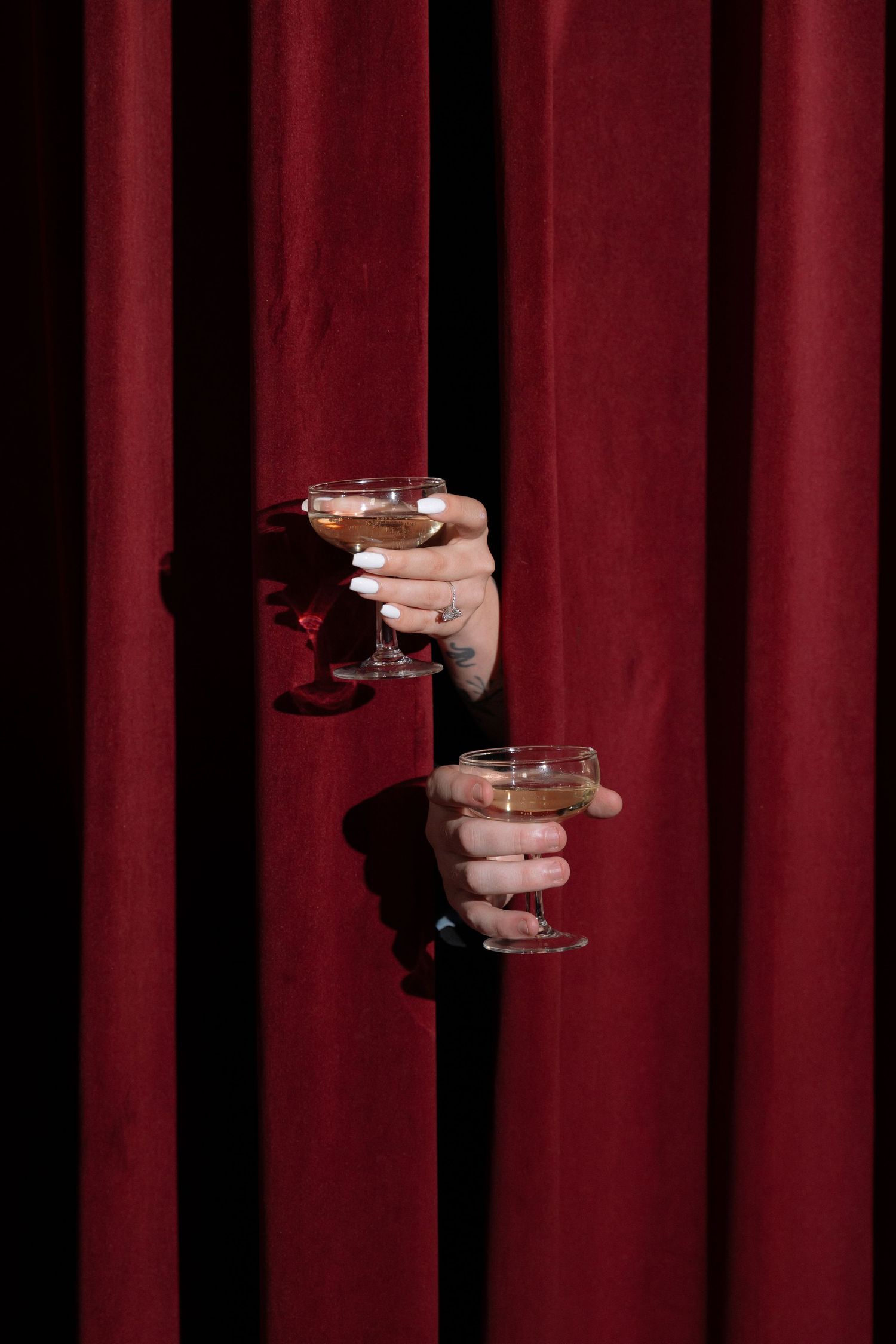 Two hands holding cocktail glasses emerging through rich red velvet curtains, with manicured nails and tattoos visible.