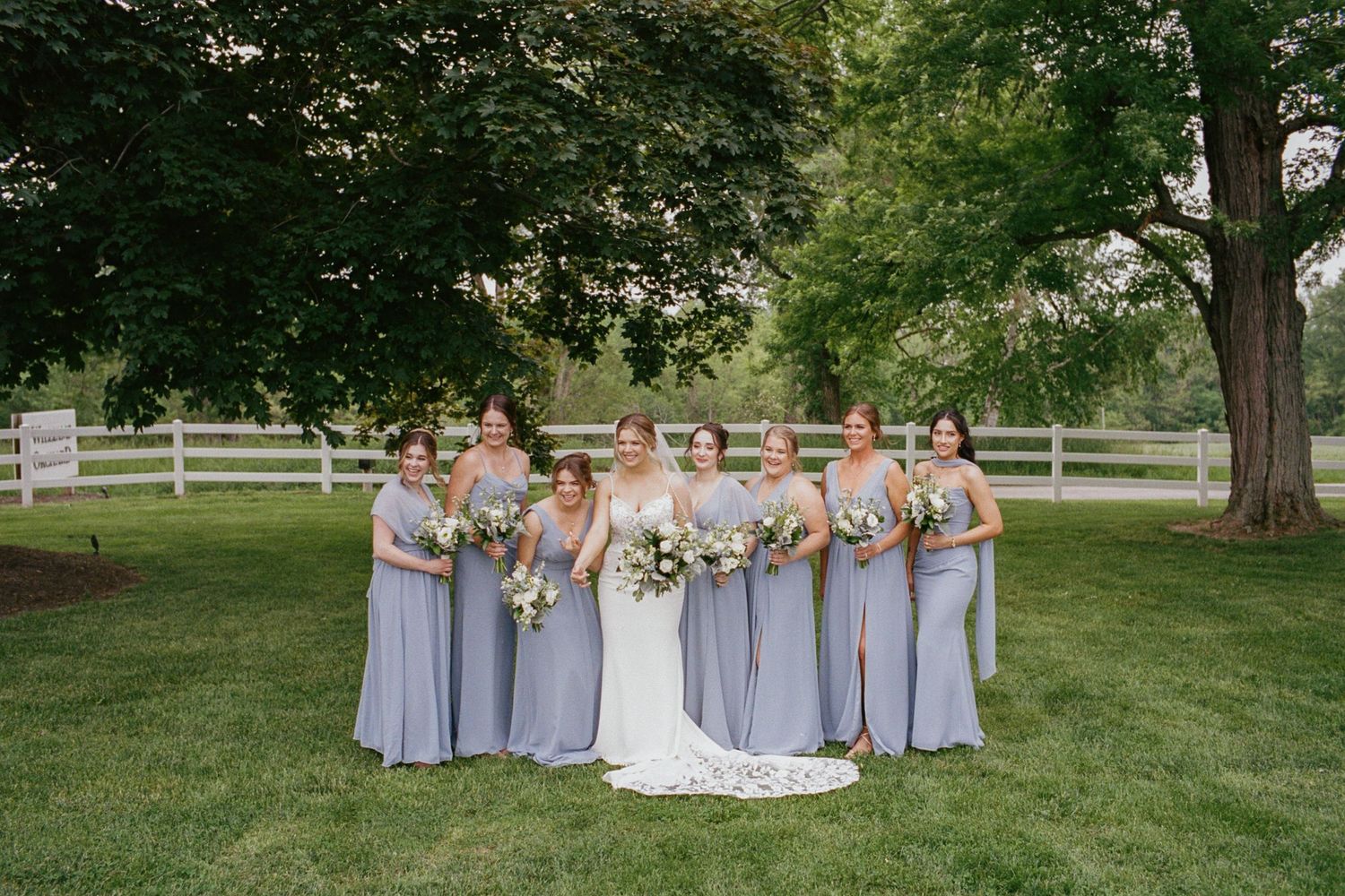 Bride in white gown poses with bridesmaids in lavender dresses holding bouquets on lush green lawn.
