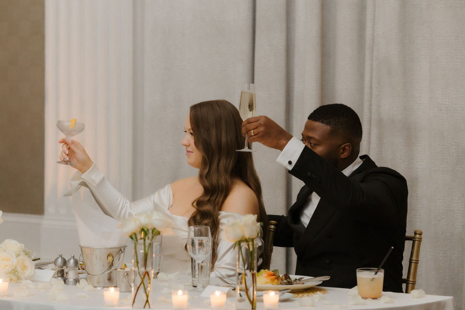 Bride and groom raising champagne glasses in a toast at their elegant wedding reception with candles.