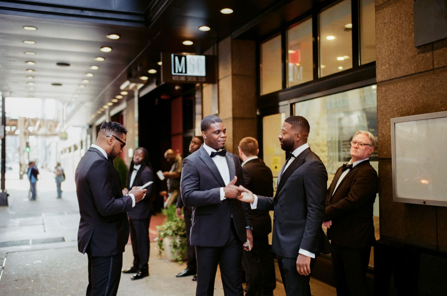 Groomsmen in tuxedos socializing outside a building before a wedding ceremony.