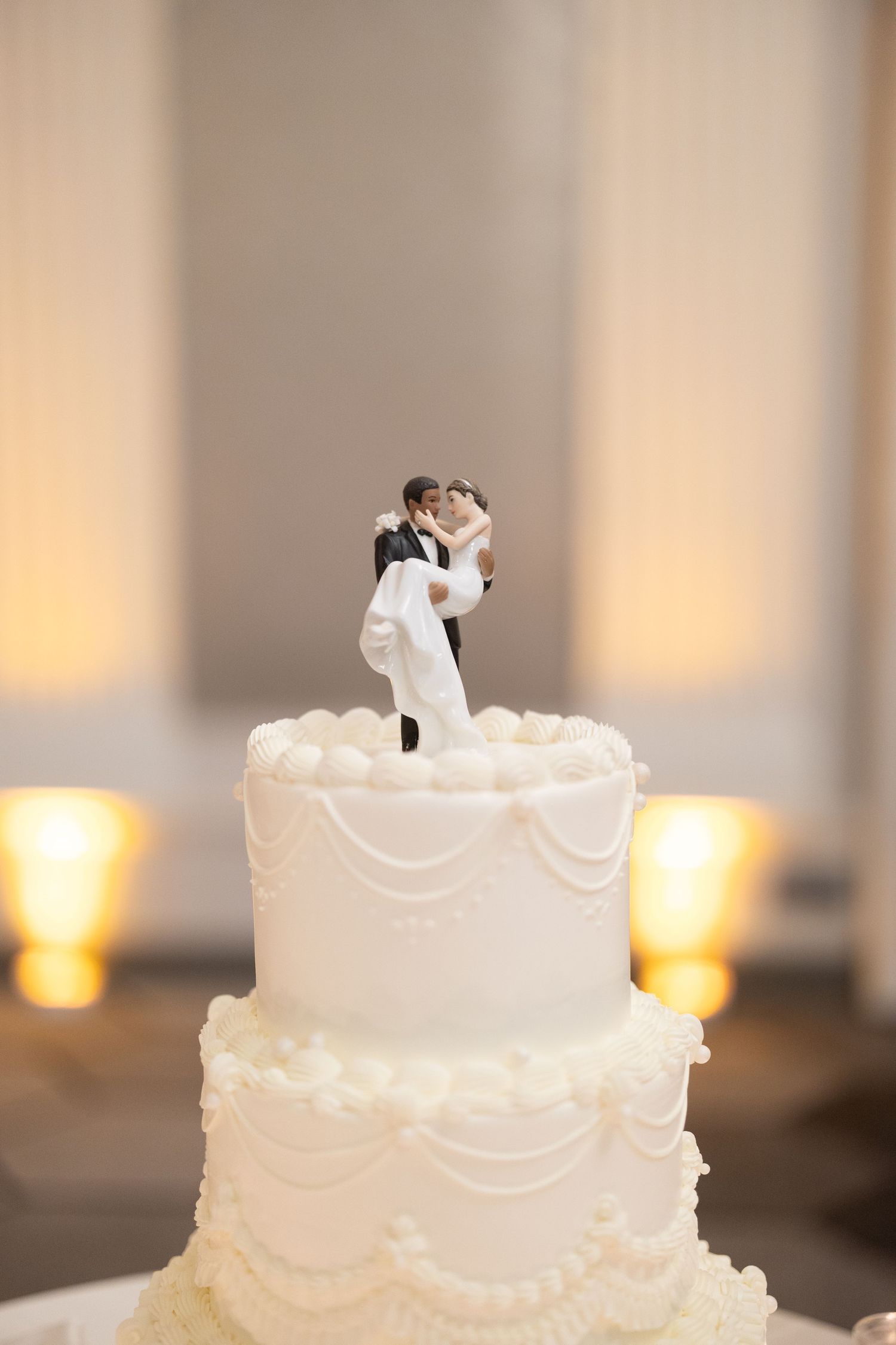 White two-tier wedding cake with classic bride and groom figurine topper, decorated with elegant piped frosting details.