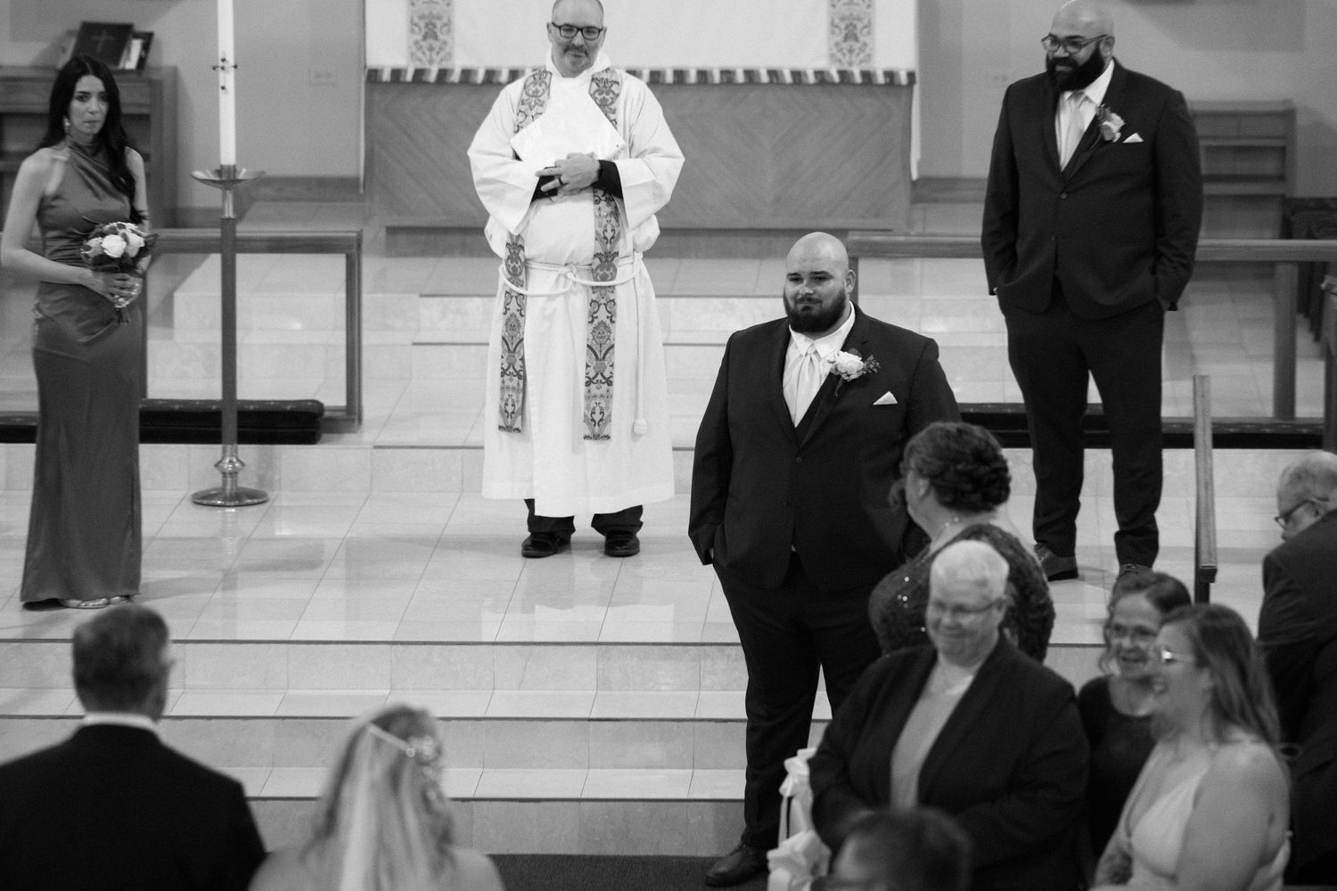 Groom stands at church altar with officiant during wedding ceremony while guests look on in black and white photo.