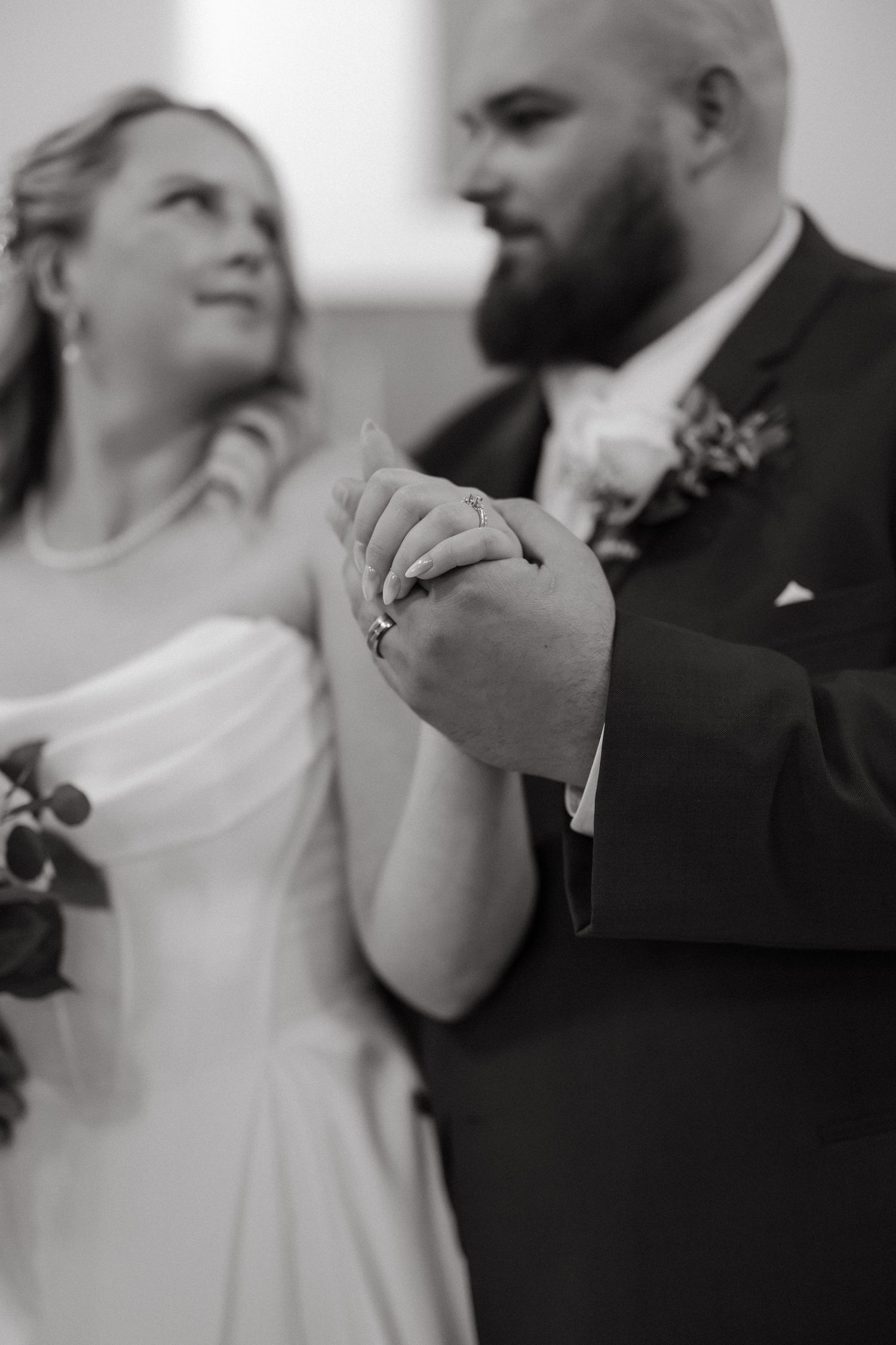 Black and white photo of bride and groom holding hands, showing wedding rings, during their wedding ceremony.