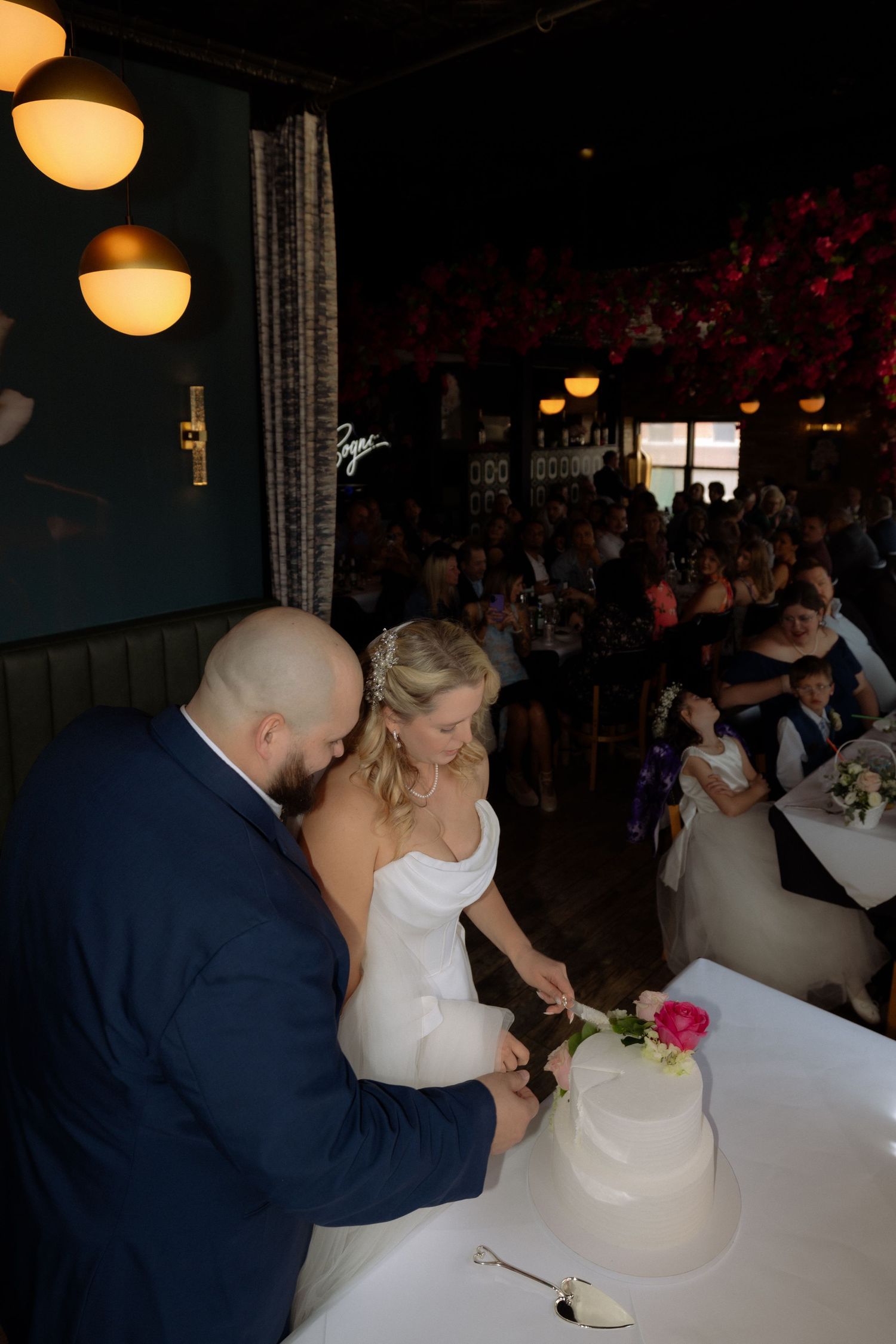 Bride and groom cutting their white two-tier wedding cake together at a reception venue filled with guests.