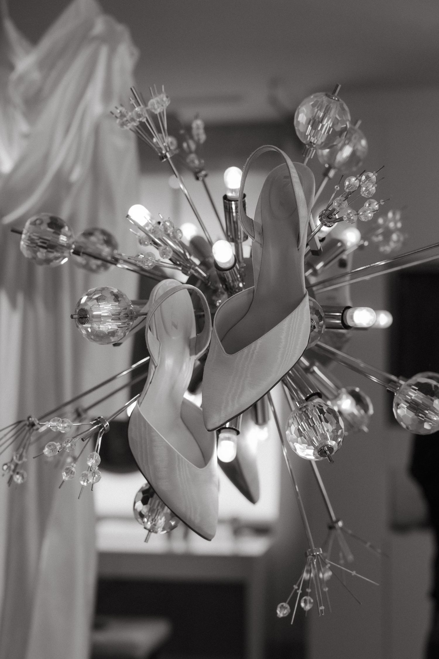 Black and white photo of elegant bridal heels displayed on a crystal chandelier with glowing bulbs.