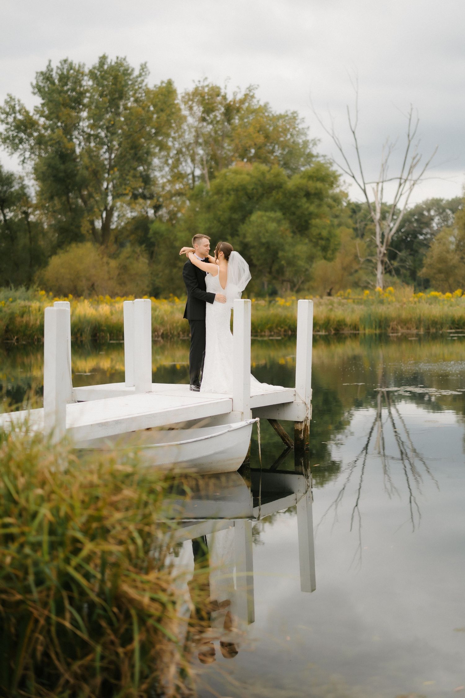 Bride and groom embracing on a white dock over a calm reflective lake surrounded by lush green trees.