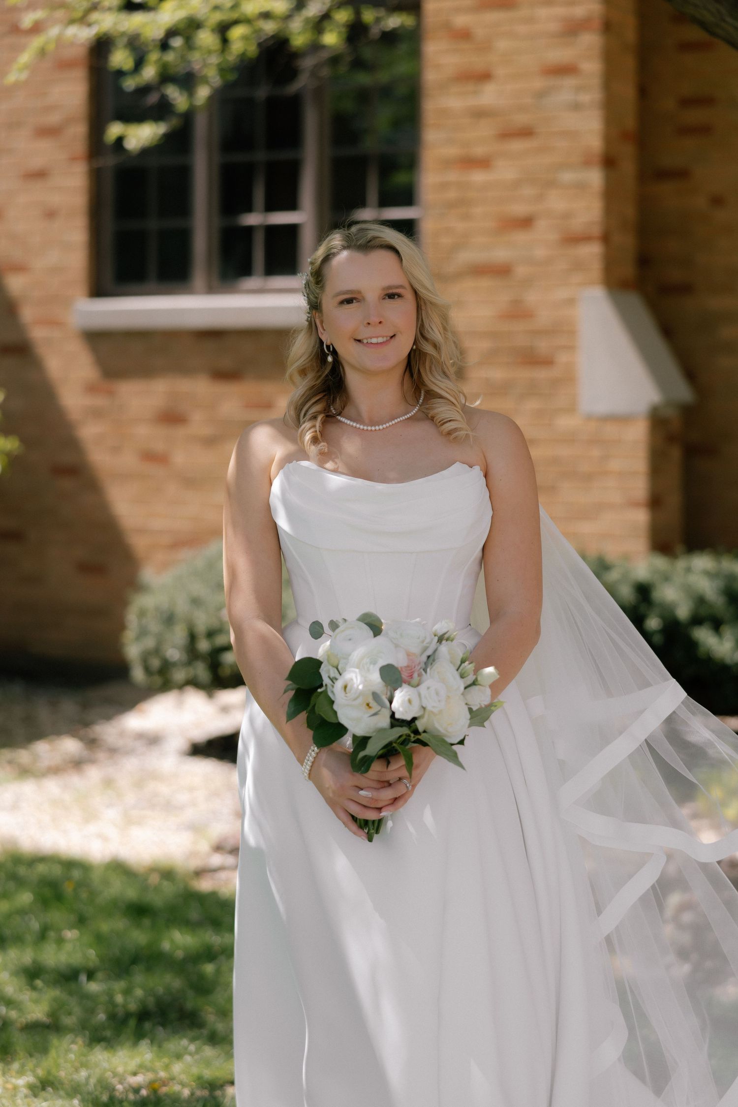 Smiling bride in white strapless gown holds white floral bouquet outdoors near brick building on sunny wedding day.