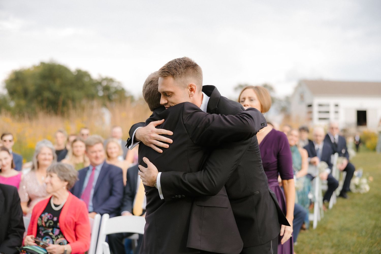 Emotional groom embraces guest during outdoor wedding ceremony with seated audience in background.
