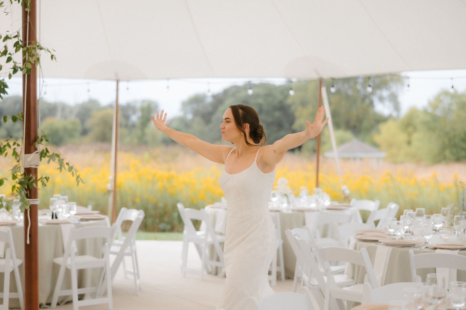 Joyful bride in lace wedding gown with arms outstretched in sunlit tent reception with floral backdrop.