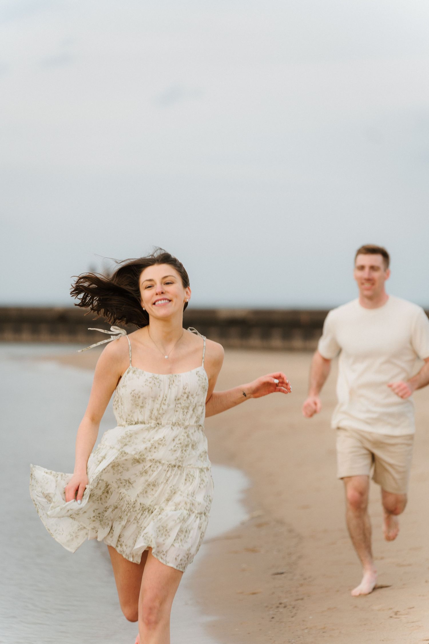 Happy couple running barefoot on a sandy beach, woman in floral sundress laughing as man chases her.
