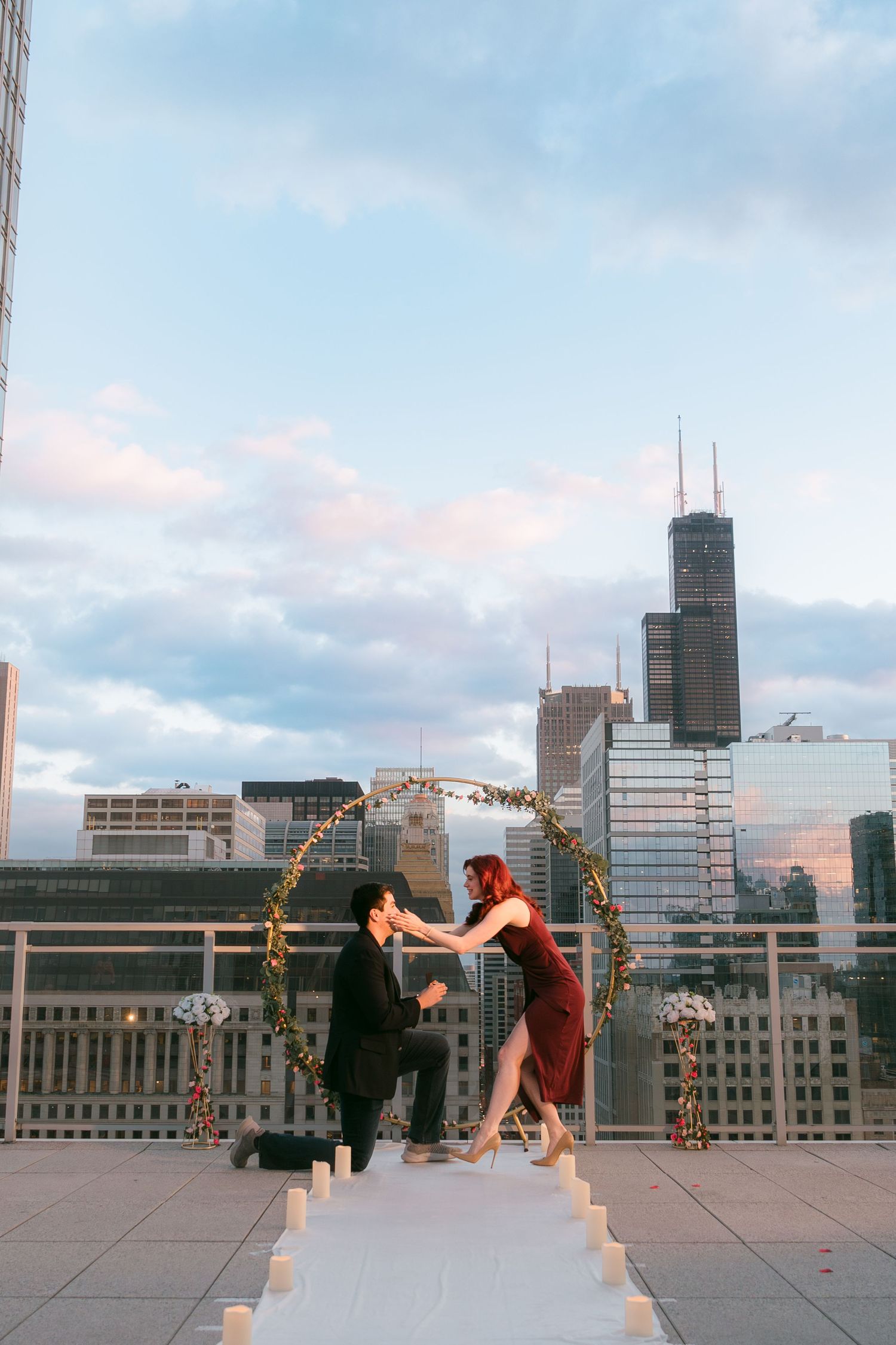 Man proposes to woman in red dress on Chicago rooftop with floral hoop arch and city skyline at sunset.