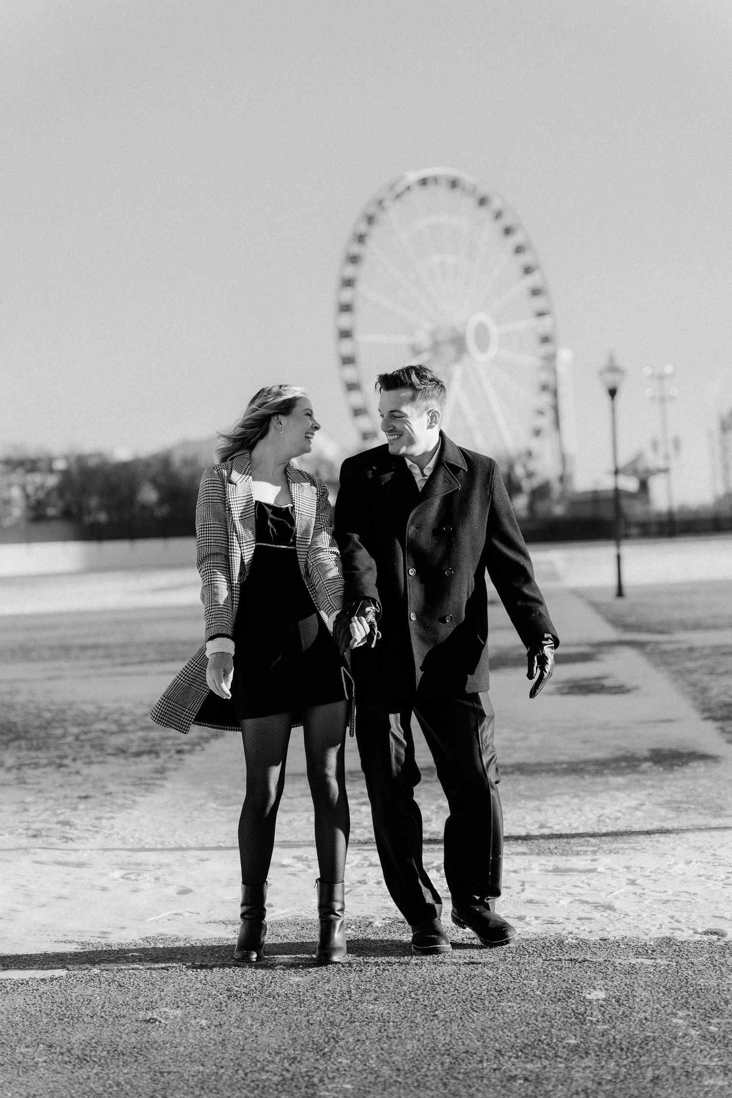 Black and white photo of a couple strolling near a Ferris wheel on a sunny winter day.