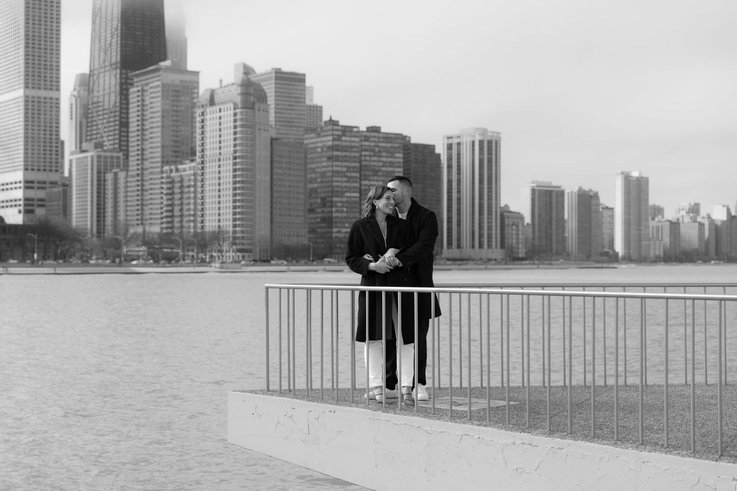 Black and white romantic photo of engaged couple embracing by Chicago lakefront with stunning city skyline.