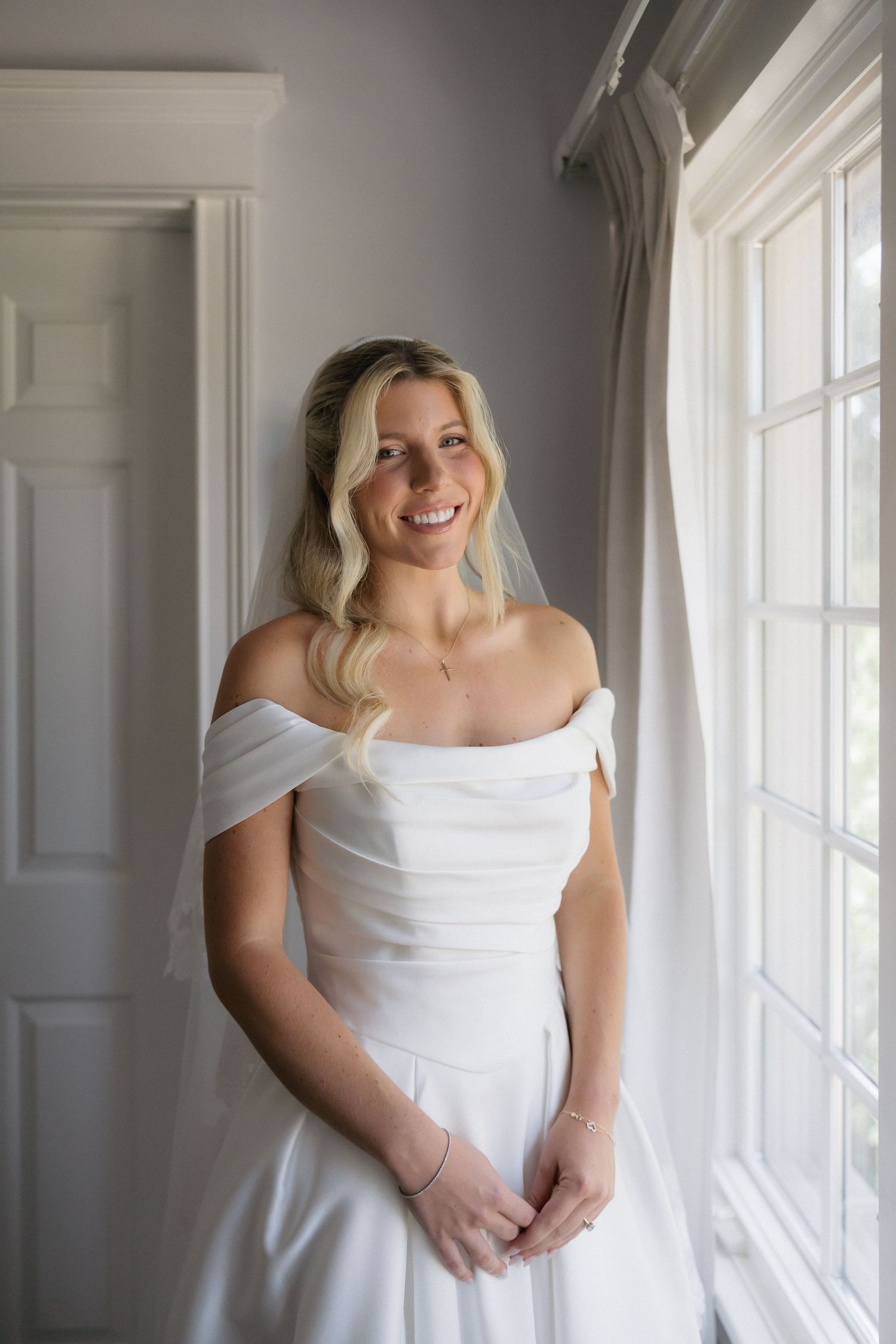 Smiling bride in off-shoulder white wedding gown stands by window in soft natural light.