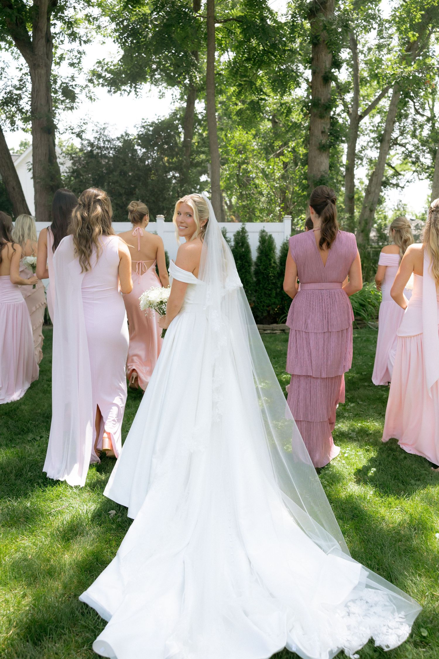 Bride in white gown and veil stands with bridesmaids in pink dresses on green lawn surrounded by trees.