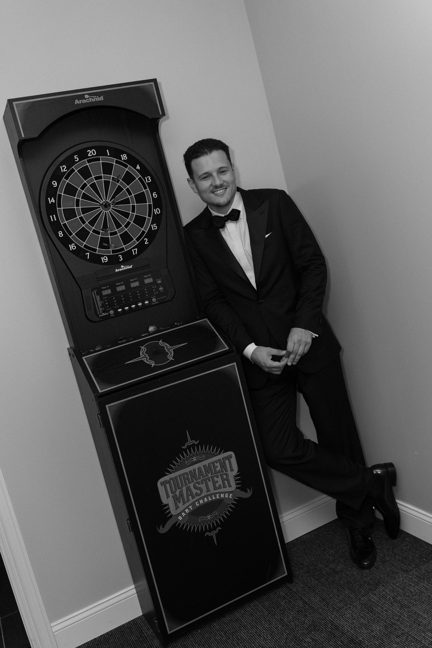 Man in tuxedo smiling beside a Tournament Master dartboard cabinet in black and white photo.