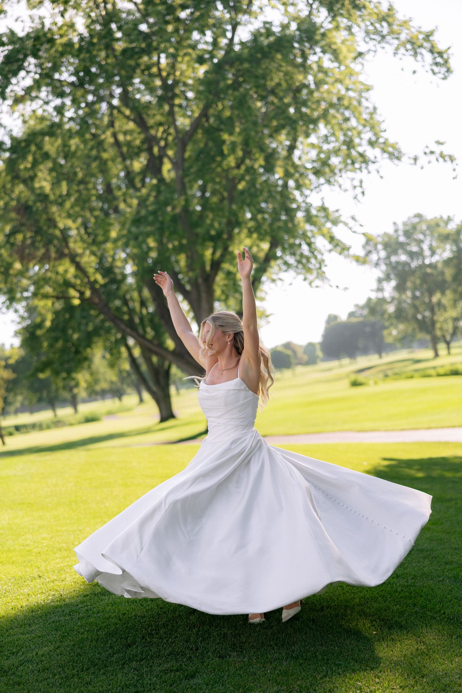 Bride in white dress twirling joyfully on lush green lawn with trees in sunny park setting.