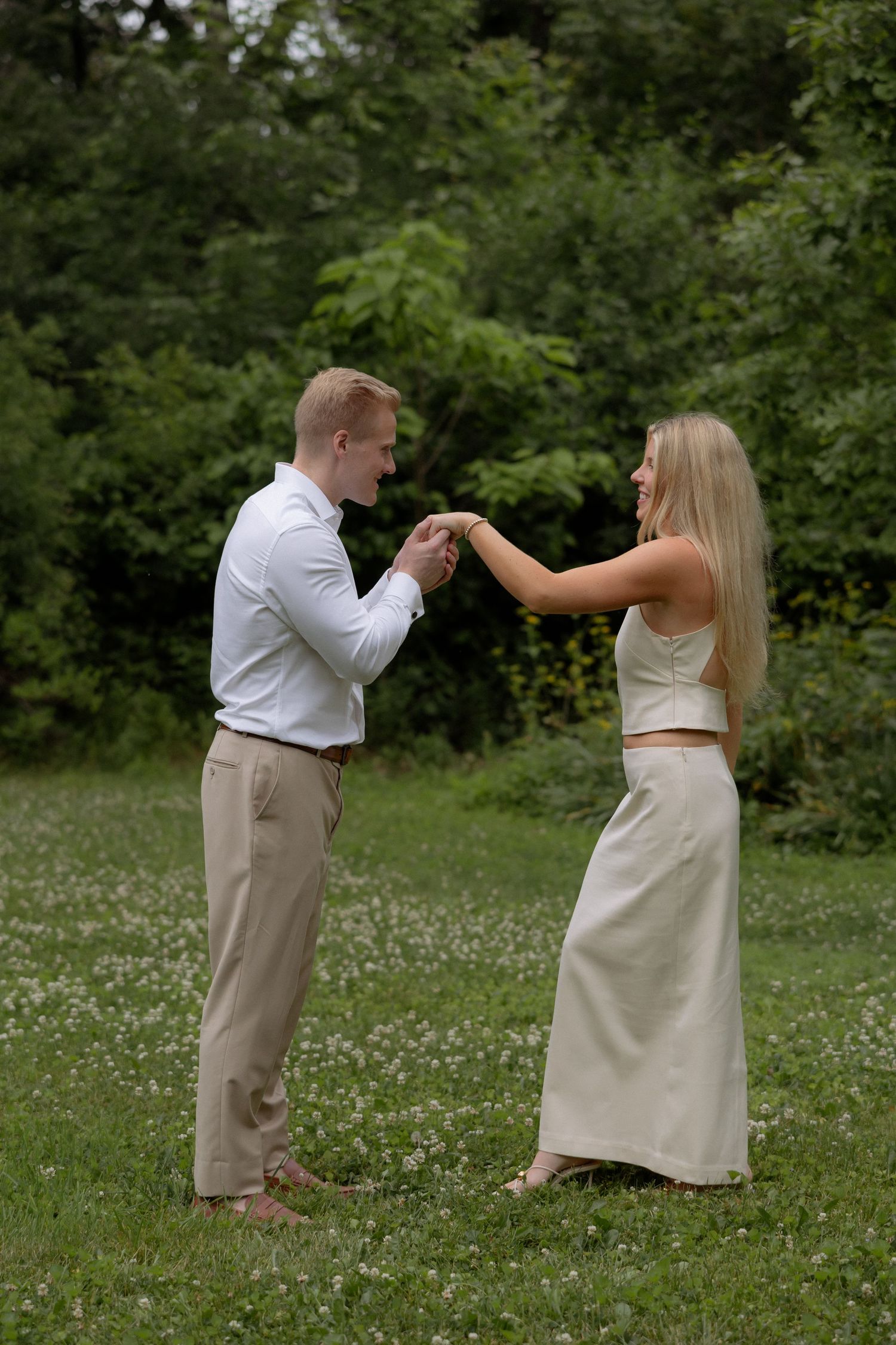 A happy couple holds hands while facing each other in a lush green meadow with wildflowers.
