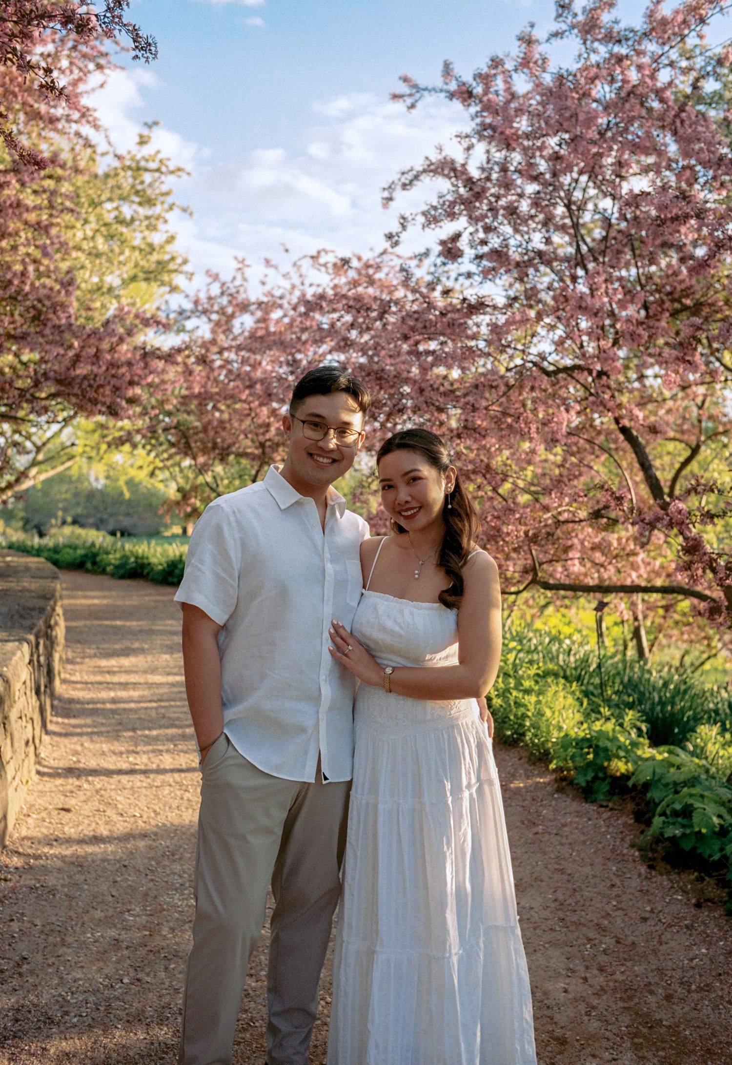 Couple posing under blooming pink cherry blossom trees on a garden path, wearing white outfits in spring.