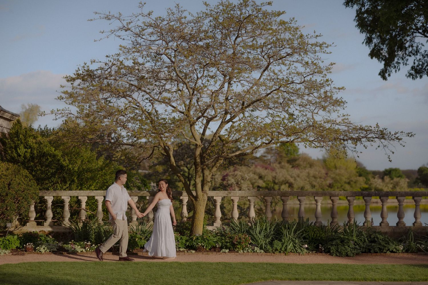Couple holding hands walking through elegant garden with stone balustrade and serene lake backdrop at golden hour.