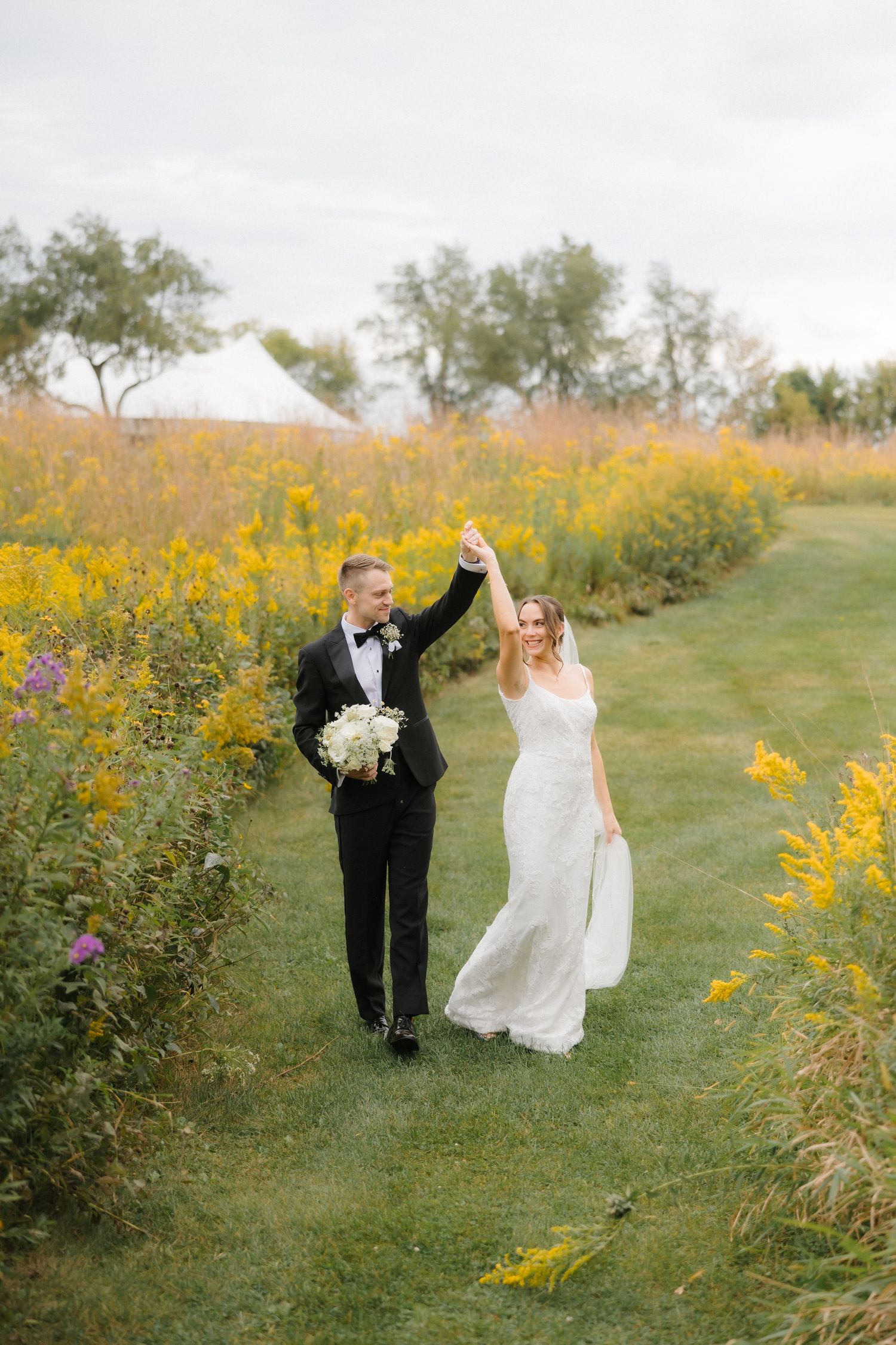Groom twirling bride along a grassy path flanked by bright yellow wildflowers with a white tent in background.