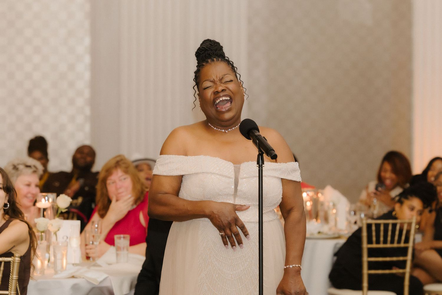 A woman in an off-shoulder white gown sings at a microphone during an elegant wedding reception.