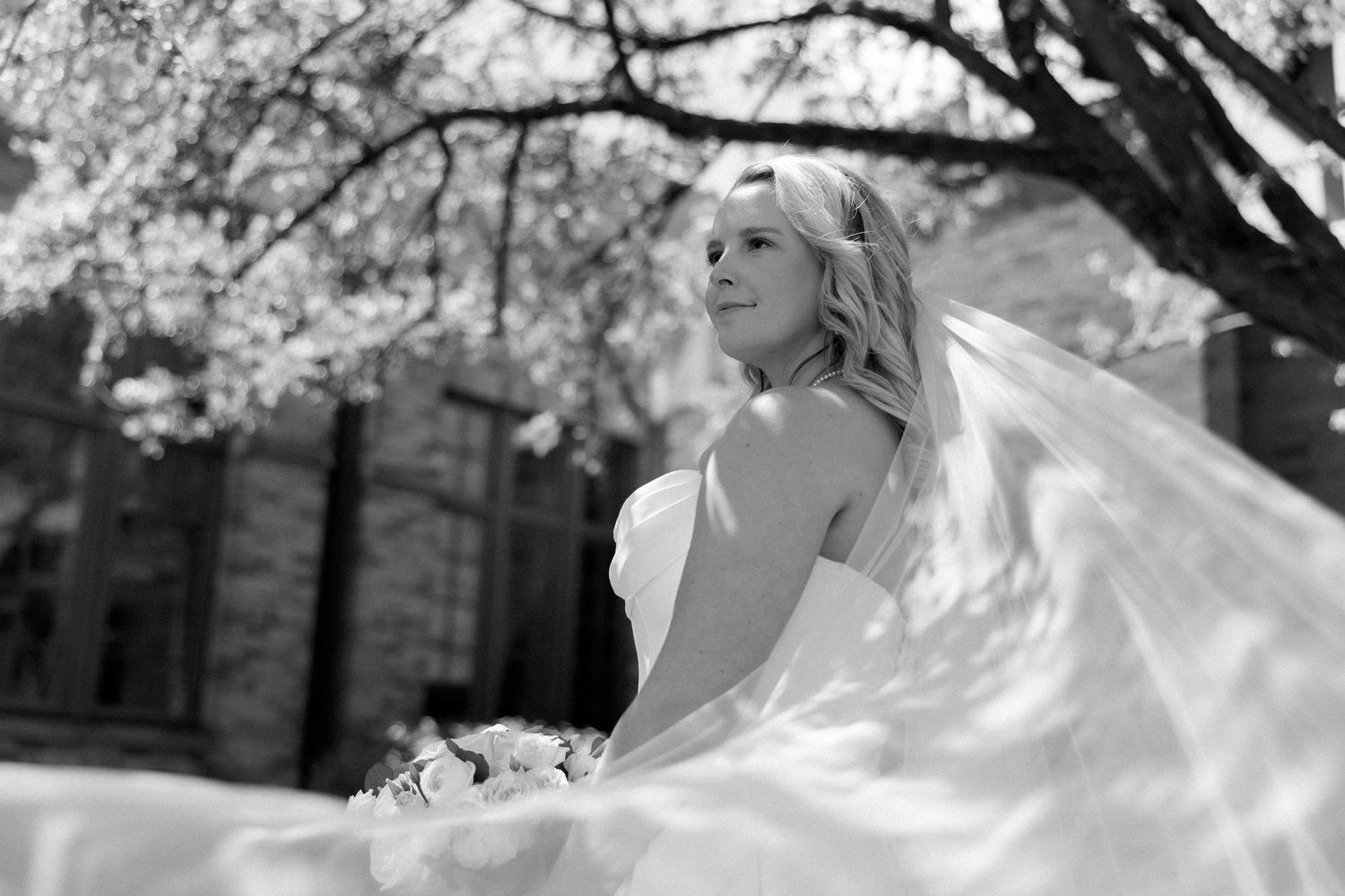 Black and white photo of a bride in a flowing white gown and veil, holding bouquet under blossoming tree.