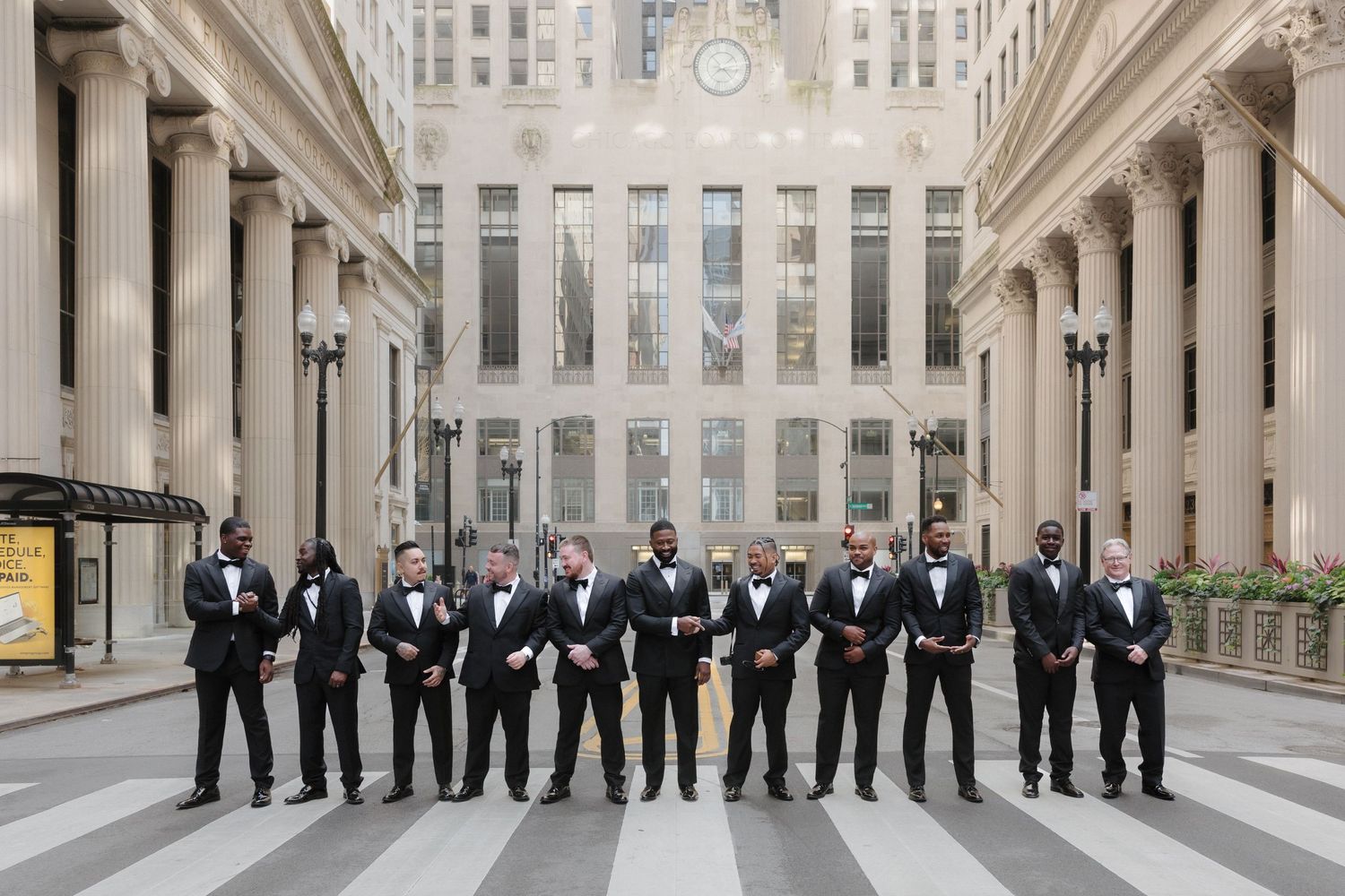 Groomsmen in black tuxedos and bow ties pose in a grand urban plaza with classical architecture.