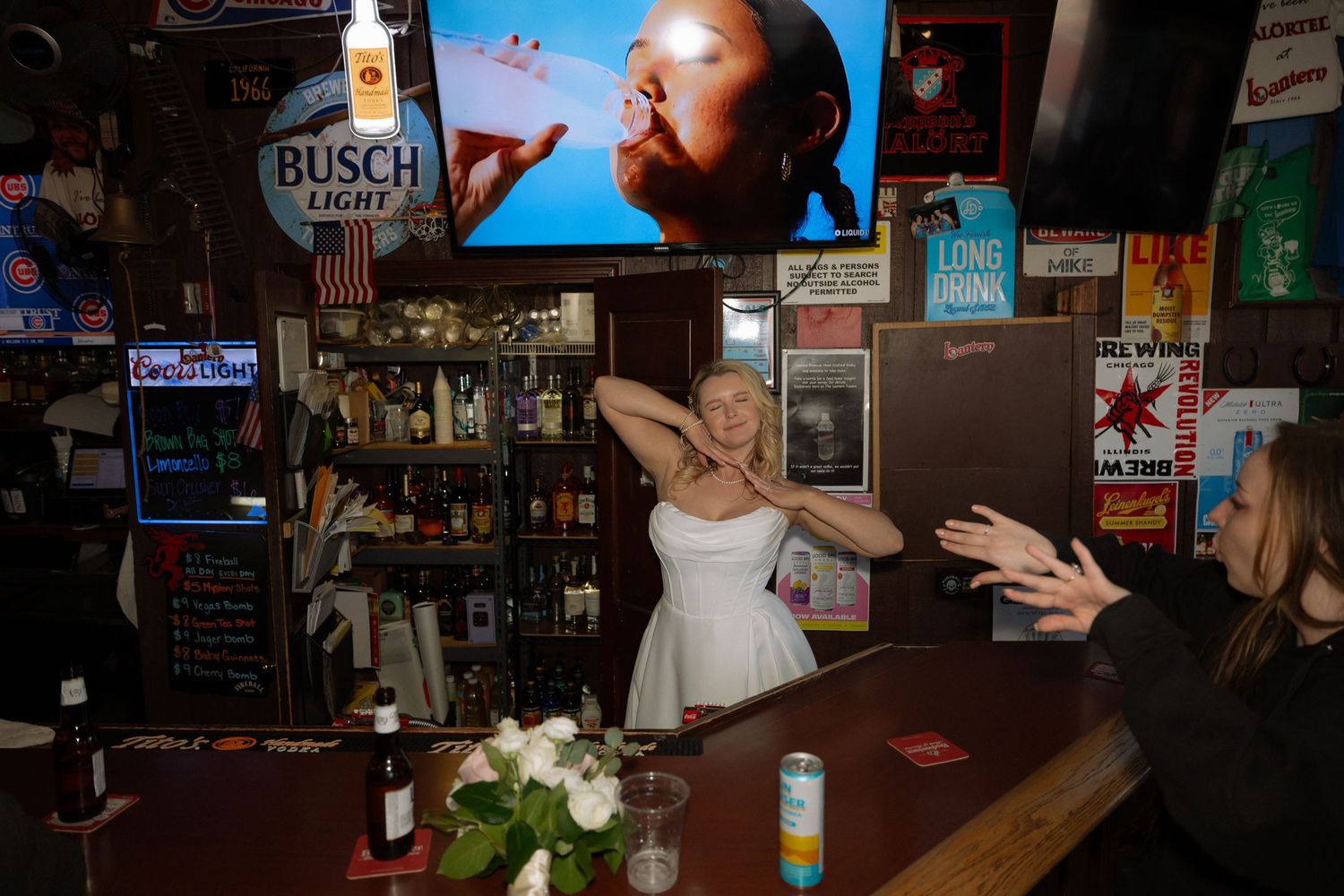 Bride in white dress dancing joyfully at a bar with Busch Light sign and colorful decor in background.