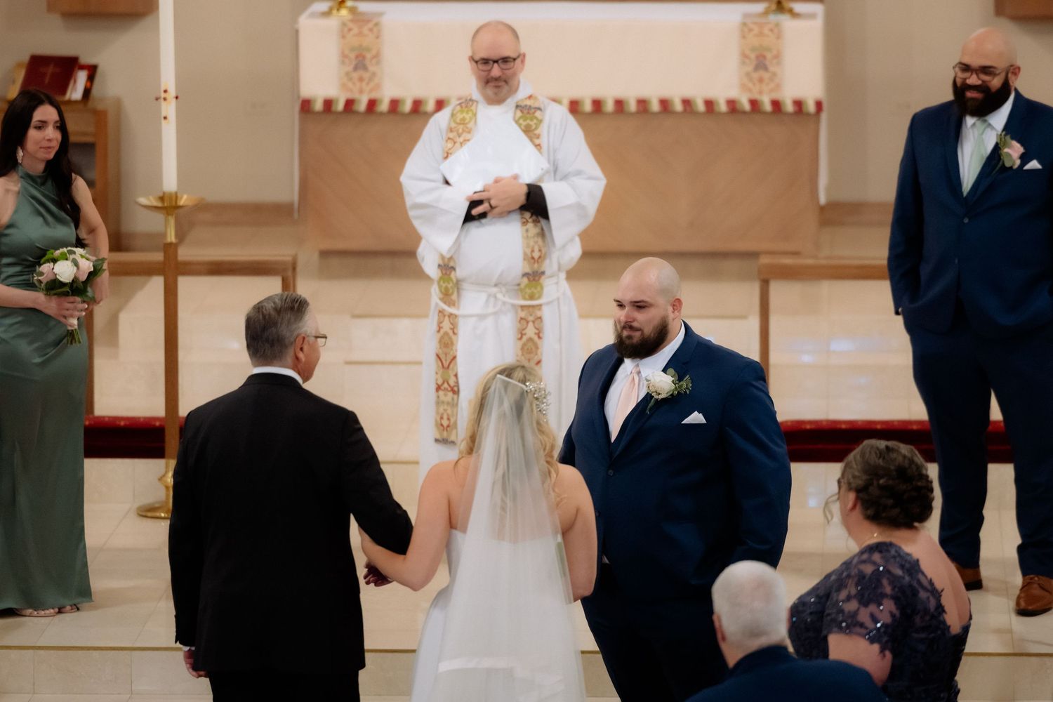 A bride and groom exchange vows at a church altar with an officiant in white and gold vestments during a wedding ceremony.