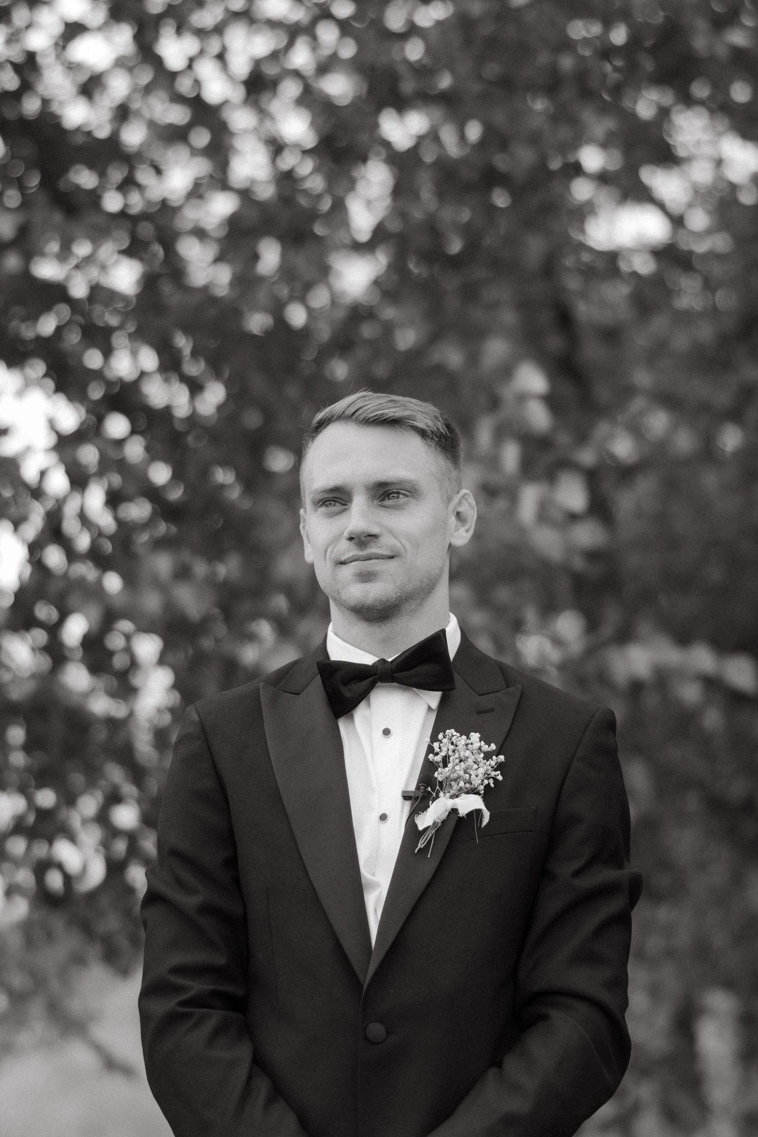Black and white portrait of handsome groom in tuxedo with boutonniere standing before leafy trees.